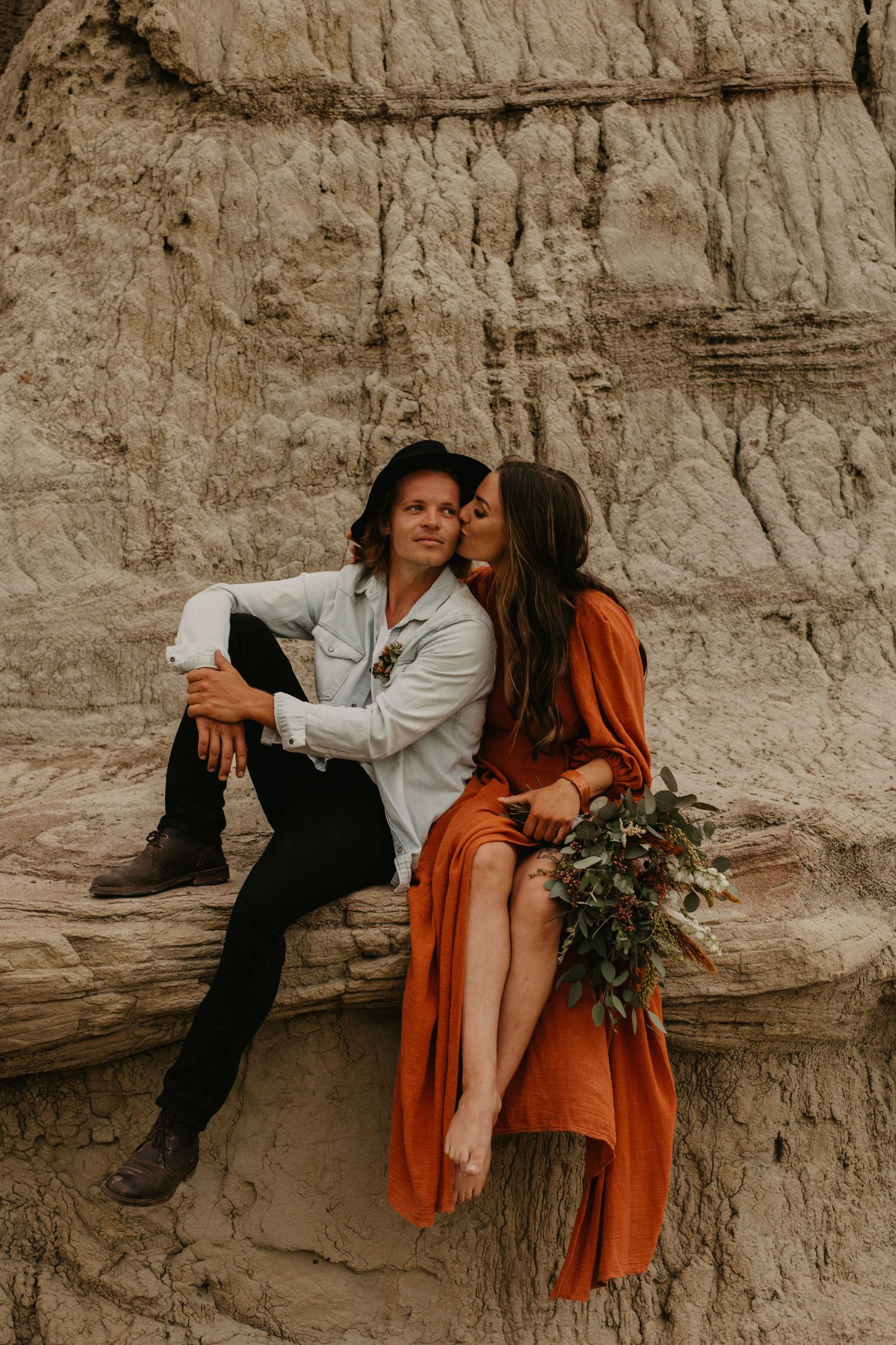 Couple in romantic pose against canyon wall, with woman in flowing orange dress holding greenery.