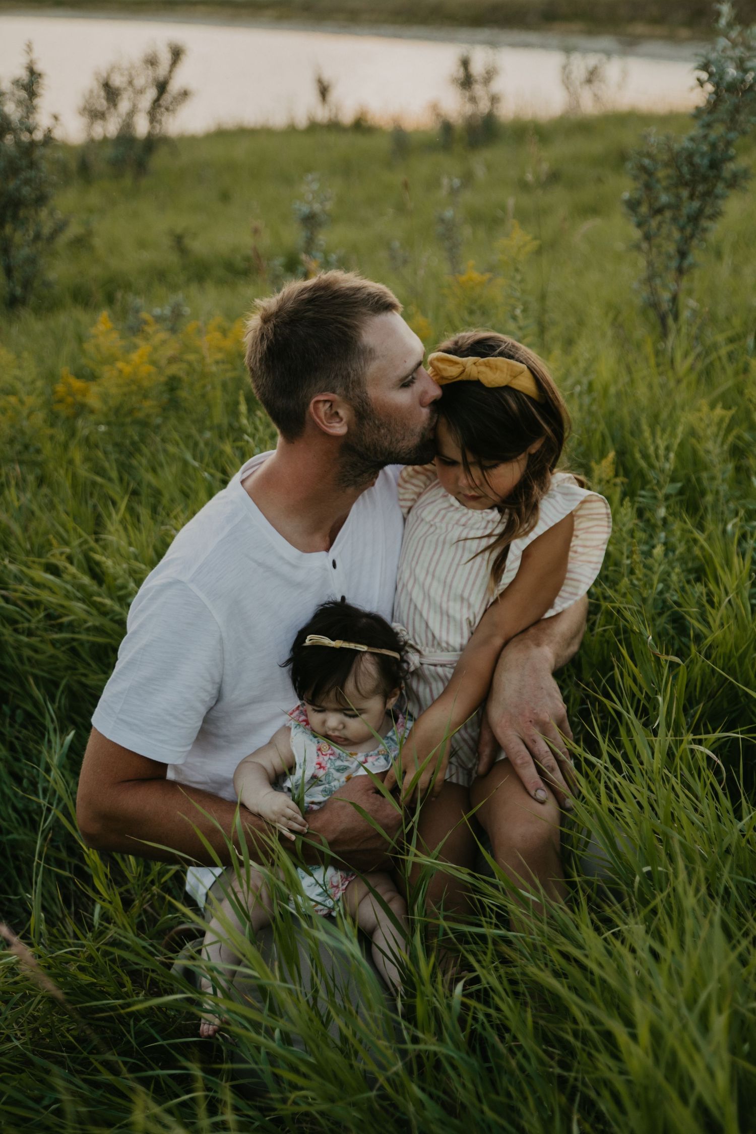 A tender family moment captured in tall grass near a lake during golden hour.