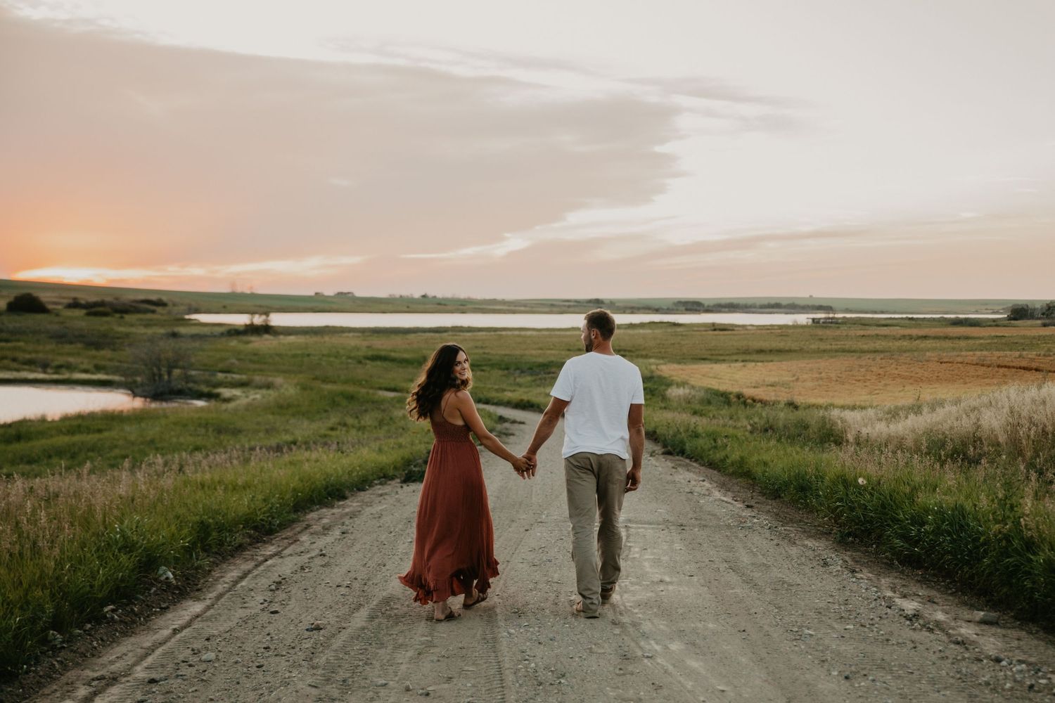 A couple walks hand in hand down a dirt road at sunset with a lake in the distance.