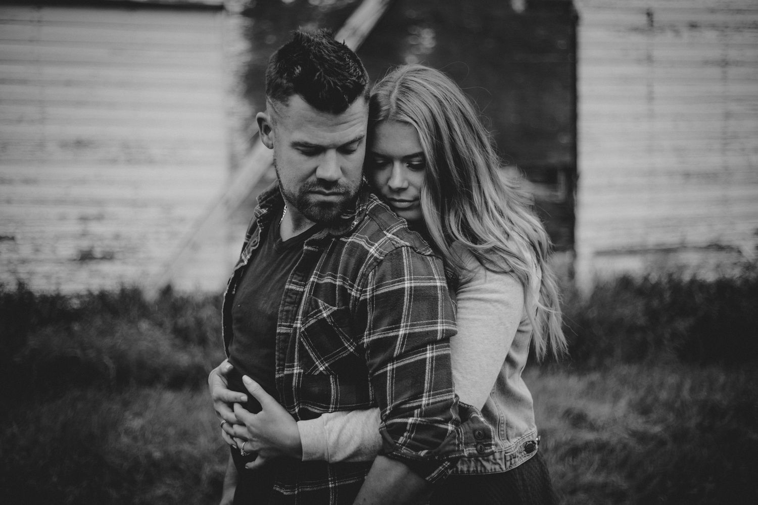 A romantic black and white portrait of a couple embracing in front of a rustic white barn.