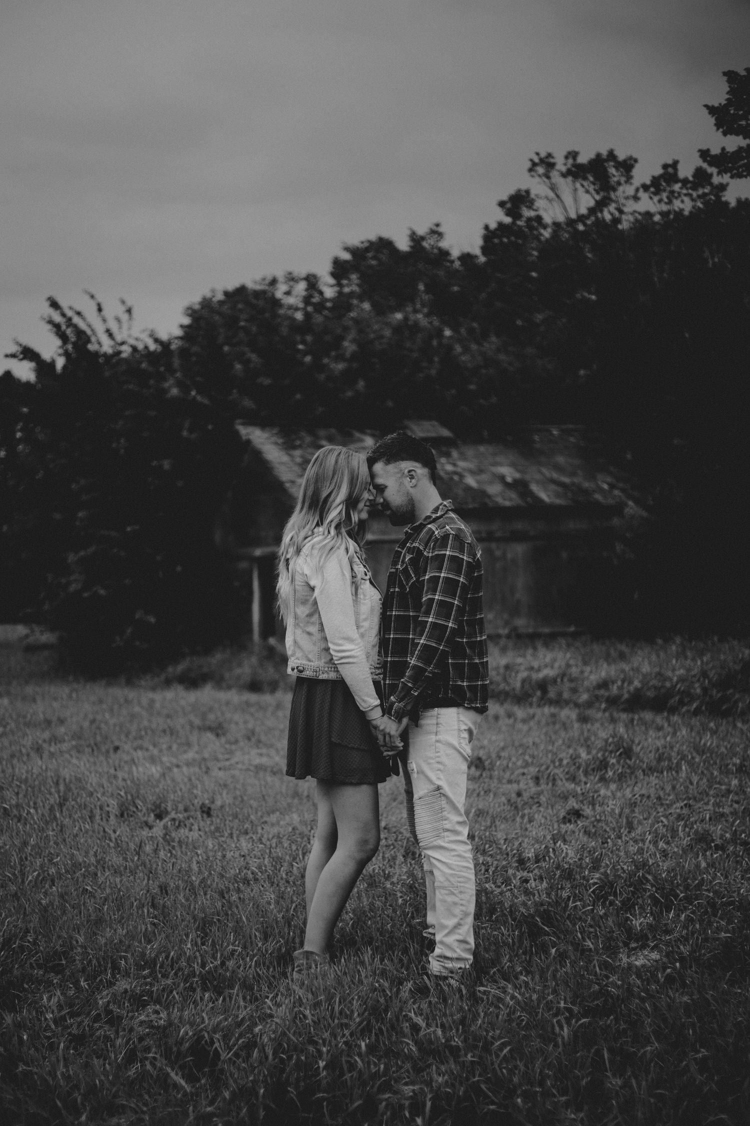 A moody black and white photograph of a couple standing close together in a rural field.