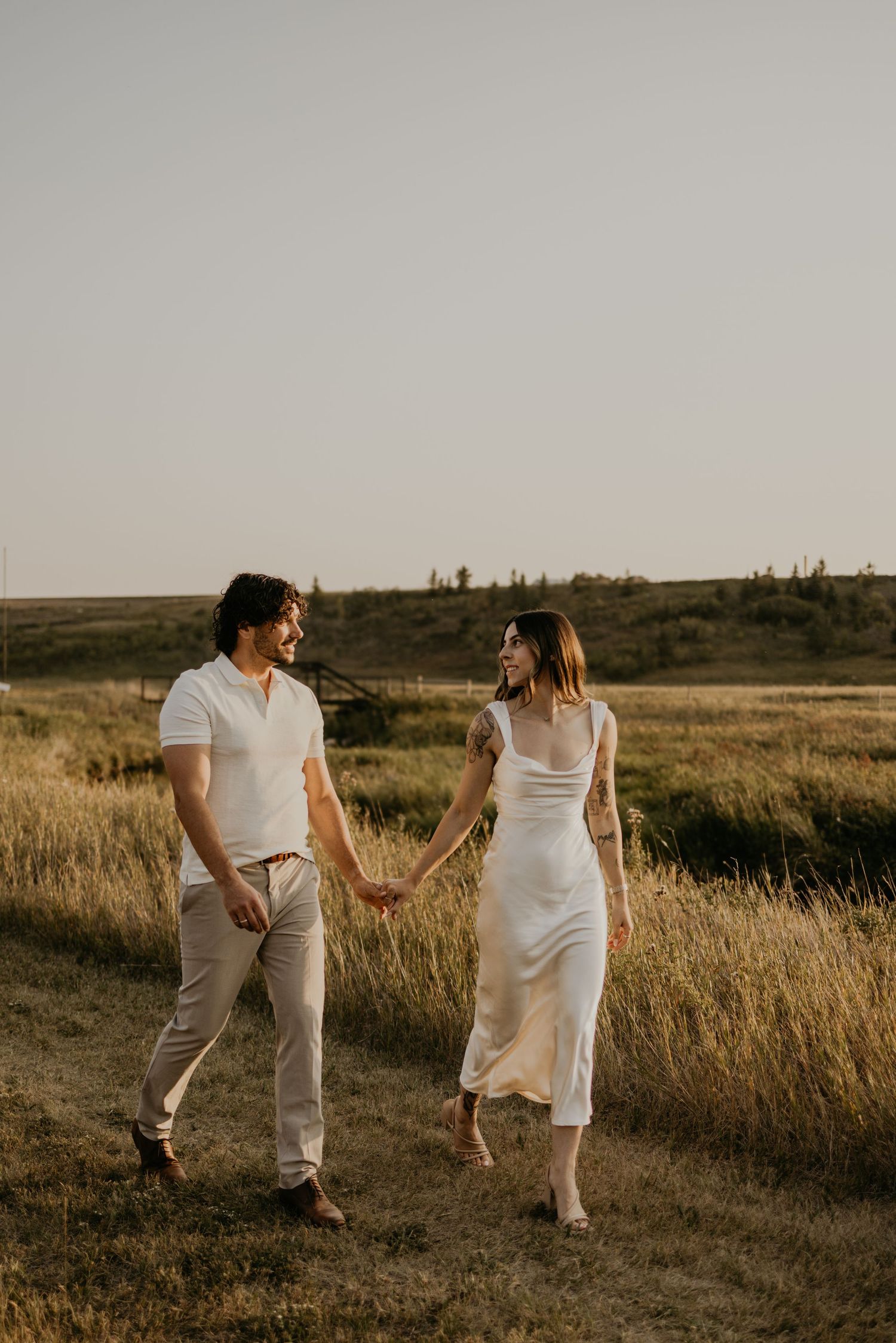 Romantic couple holding hands while strolling through rustic countryside meadow at dusk.
