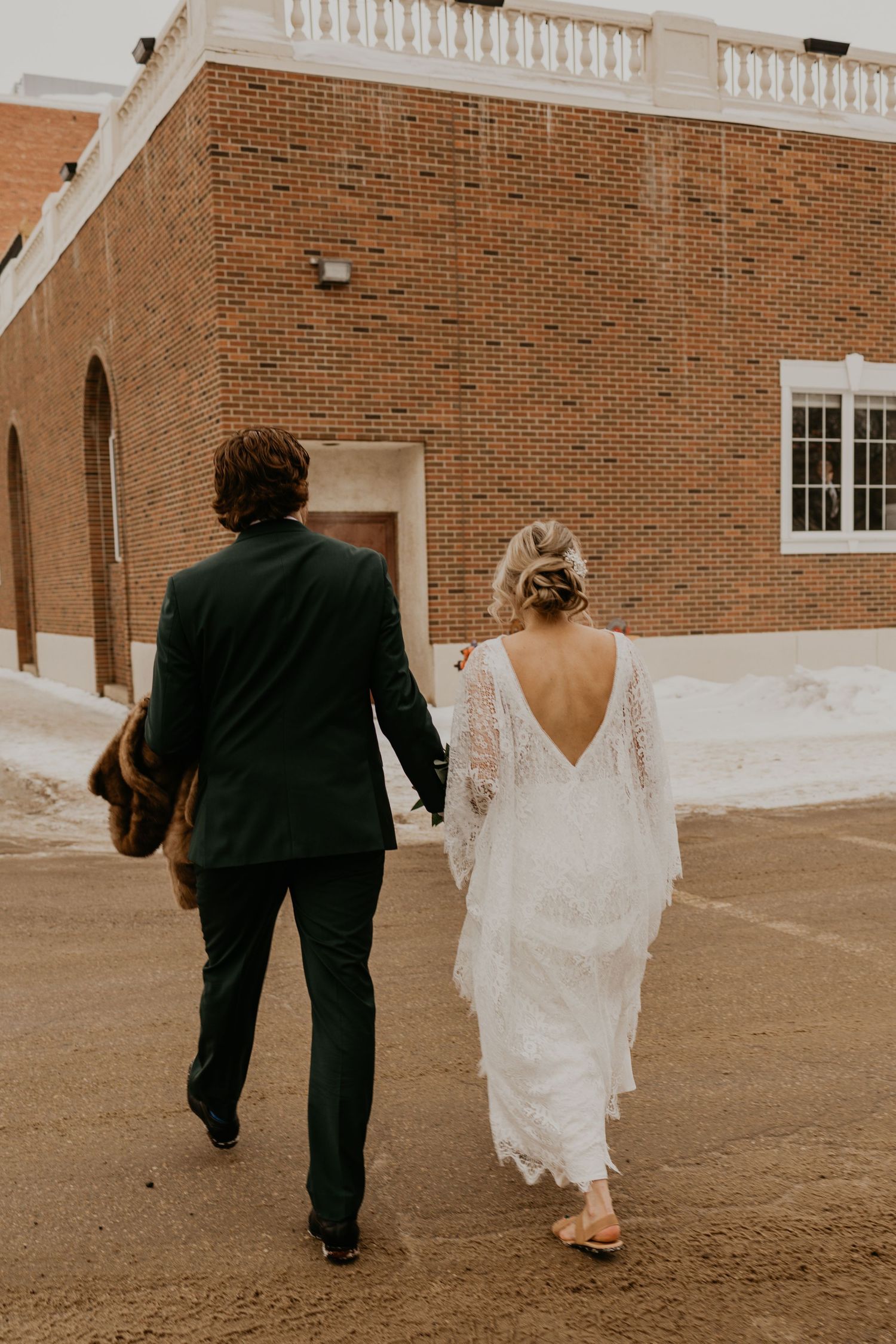 A couple walks hand in hand with a brick building in the background.