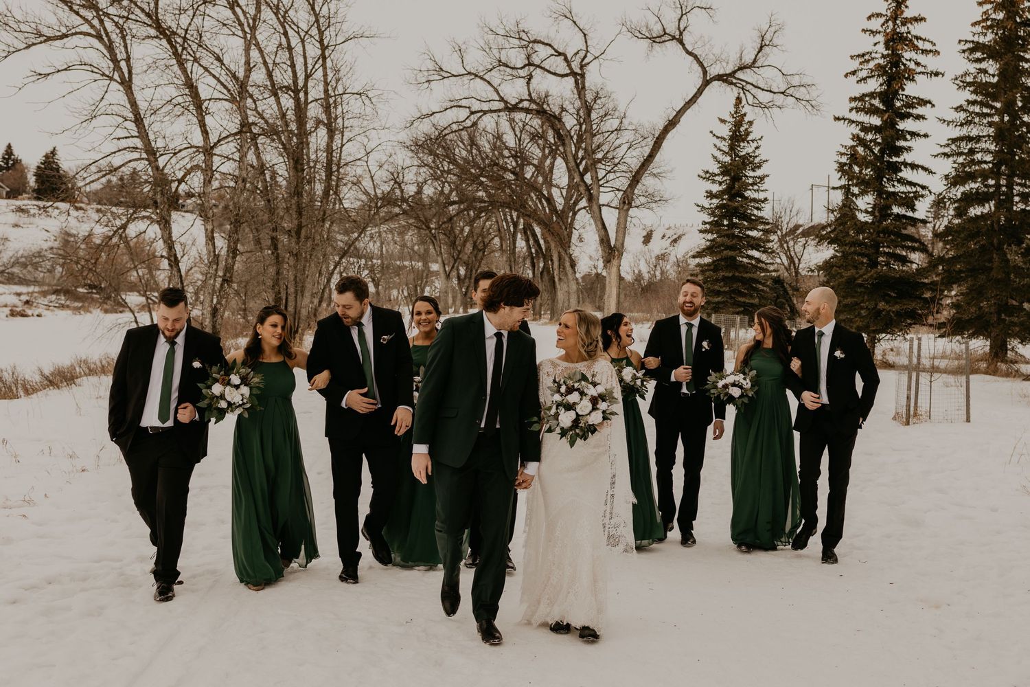 Wedding party poses while walking in snow with bridesmaids wearing dark green dresses and groomsmen in black suits.