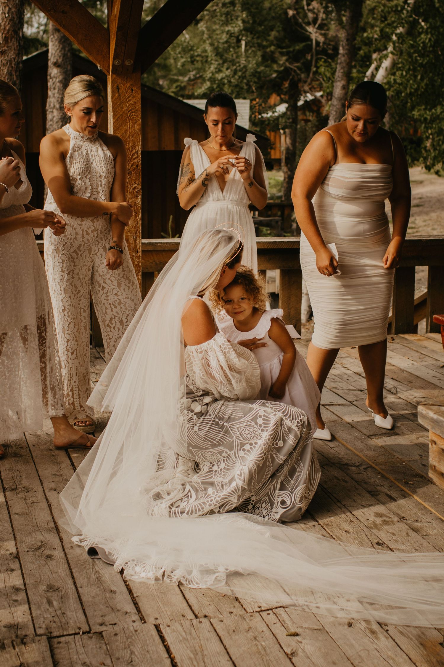 Wedding party members help adjust the bride's long white veil on a wooden deck.
