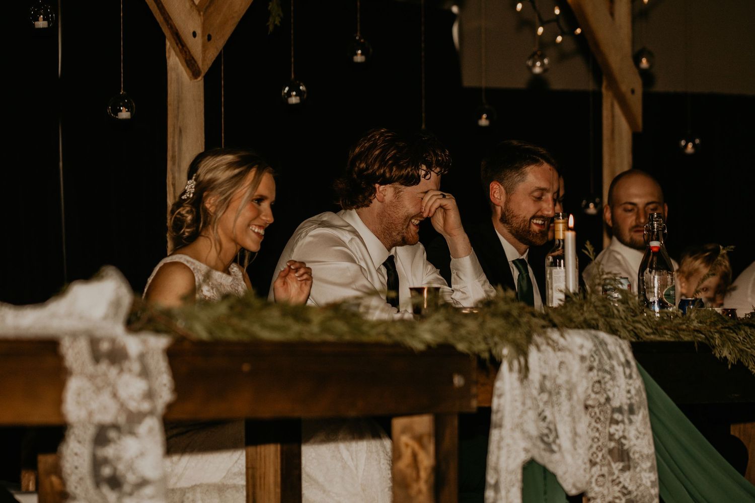 Wedding party seated at rustic wooden head table during reception dinner.