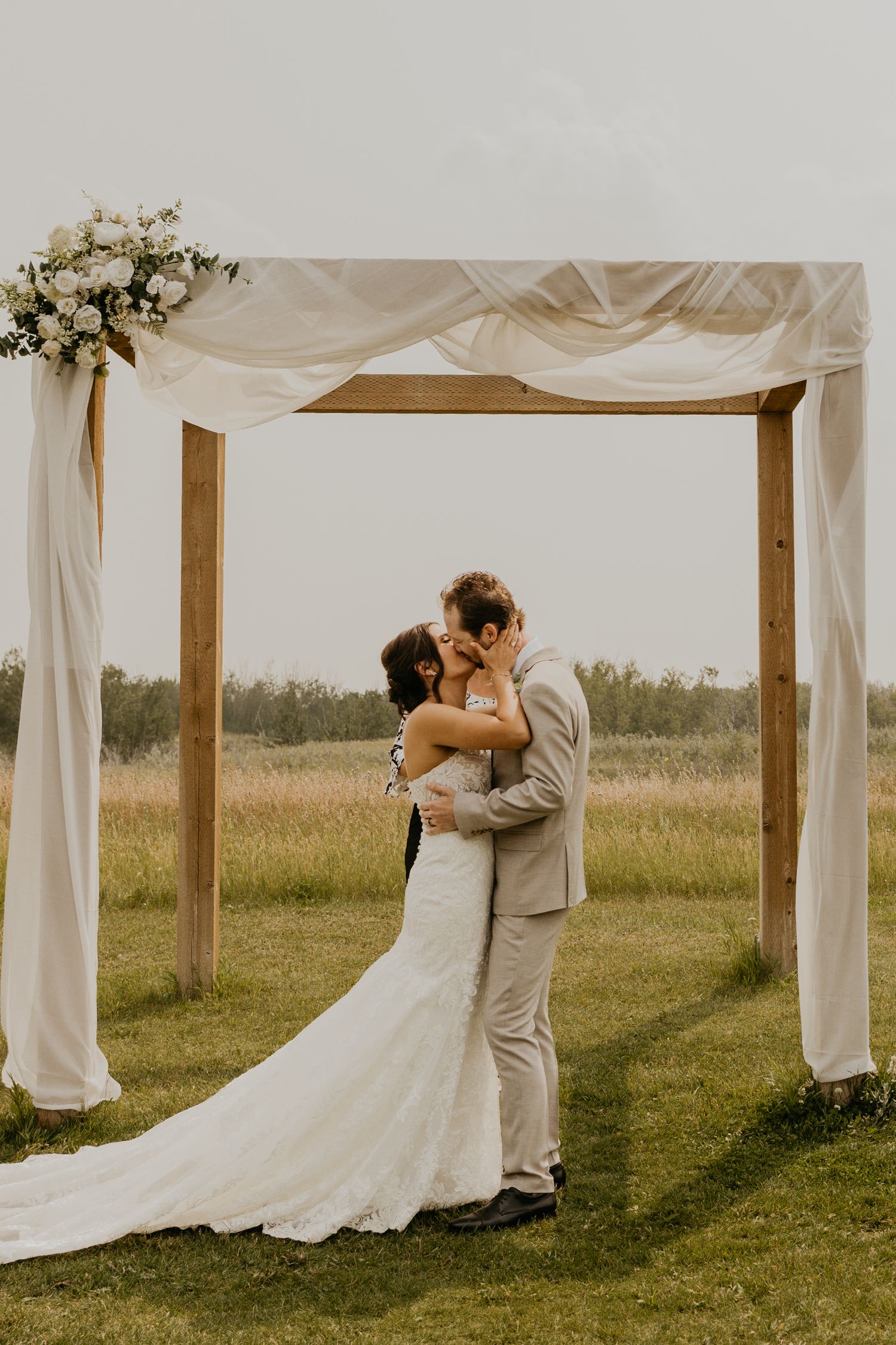 Romantic ceremony moment under wooden wedding arch draped with white fabric in outdoor field setting.