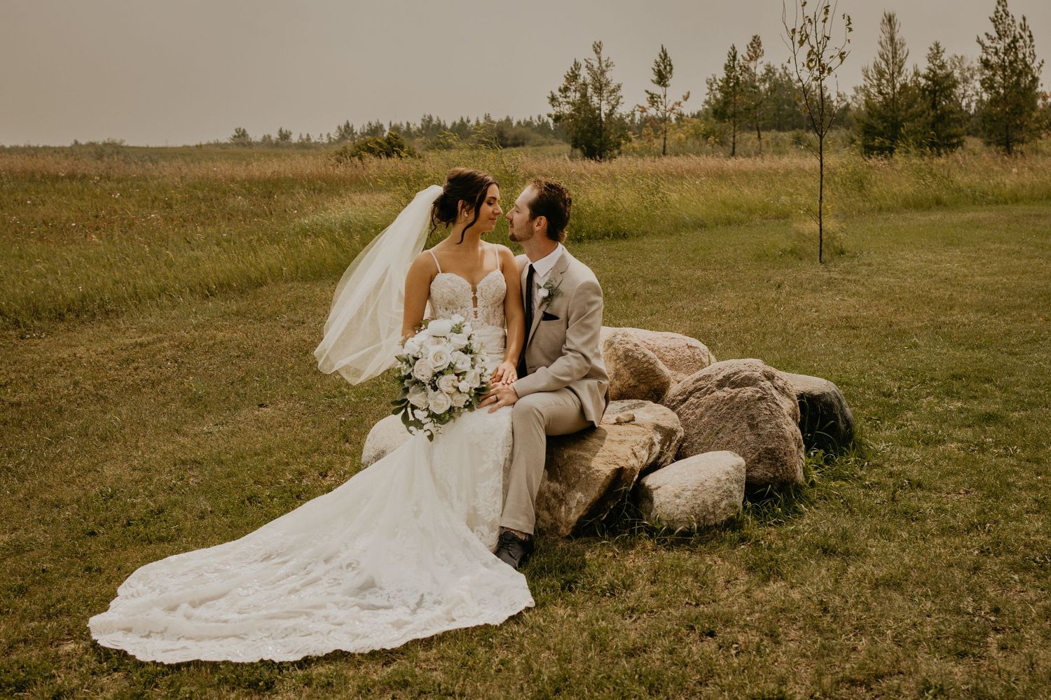 Couple shares intimate moment on stone bench with bride's dress flowing across natural landscape.