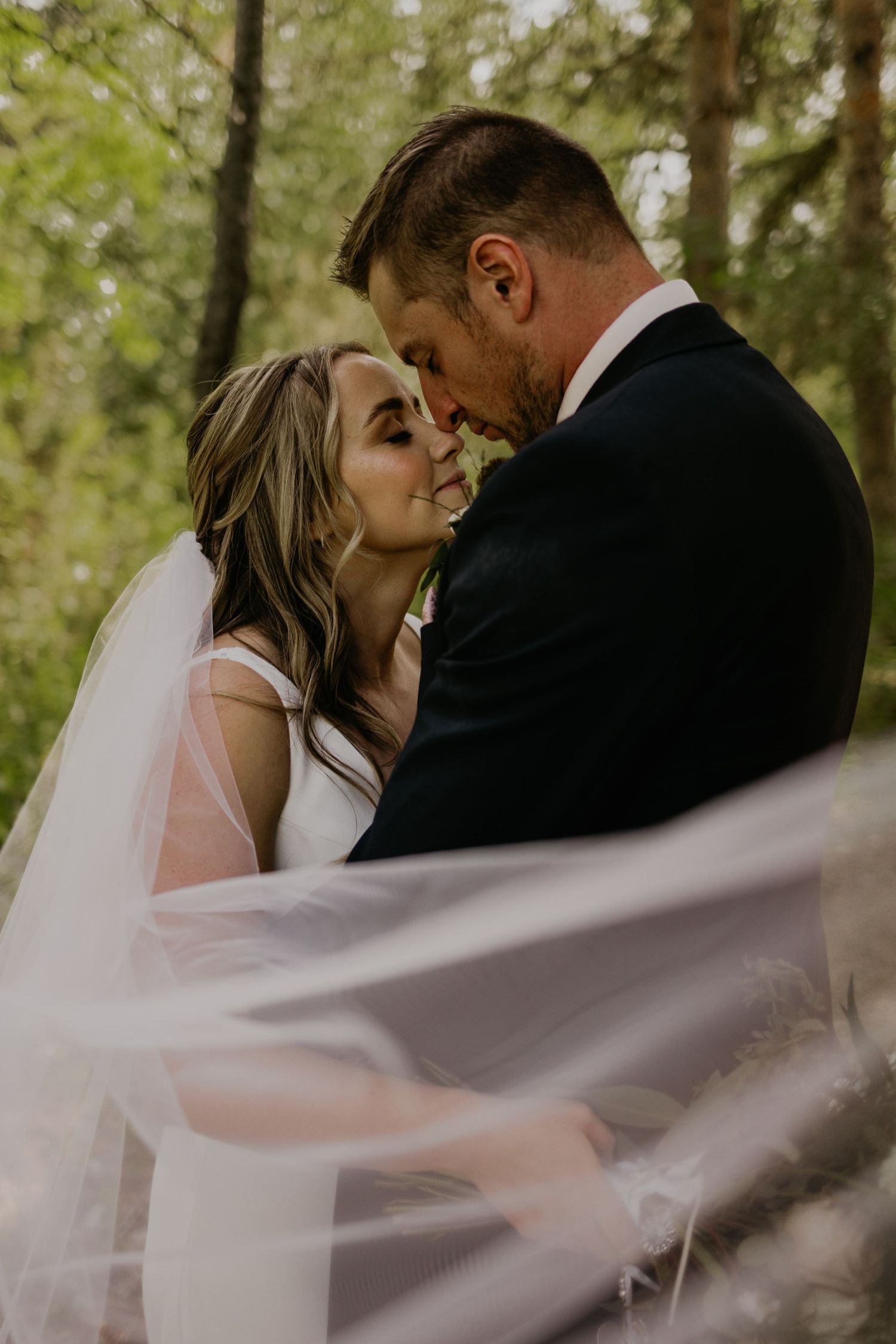 A tender moment between newlyweds with flowing veil in golden forest light.