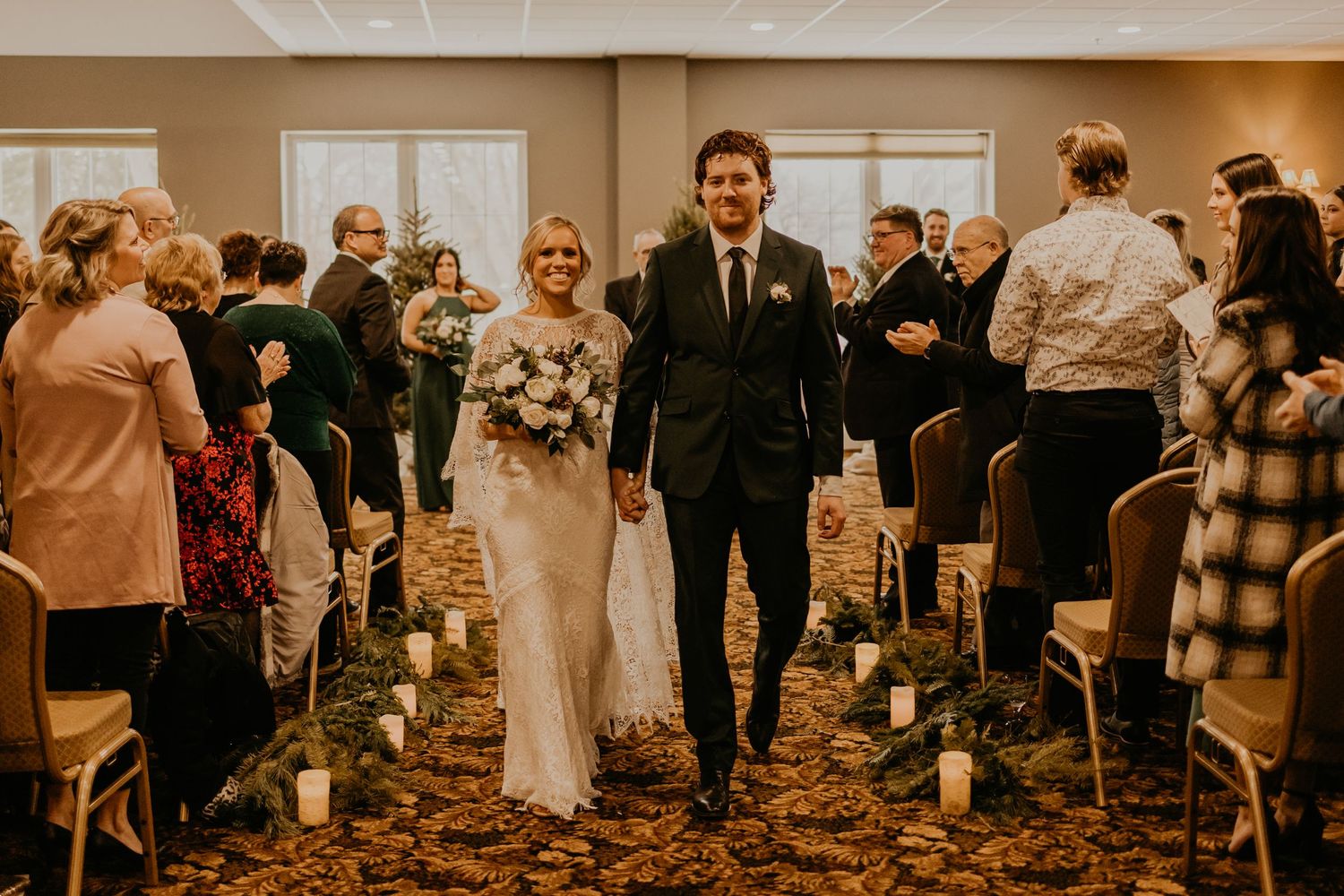 Newlyweds walk down the aisle while guests celebrate in a warm indoor ceremony space decorated for winter.