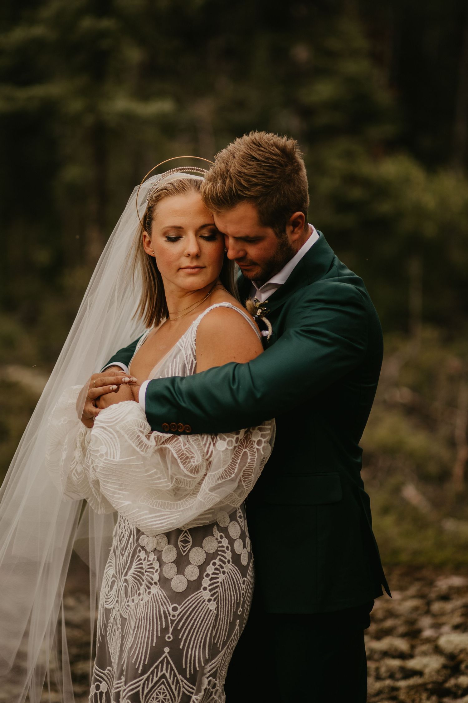 Romantic wedding portrait with emerald suit and lace dress in moody forest setting.