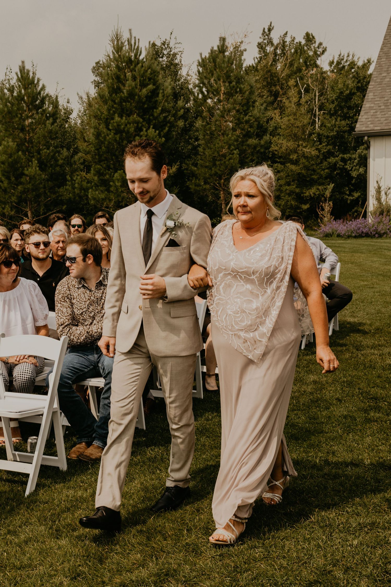 A processional scene at a rustic outdoor wedding with attendees walking across a grassy lawn wearing formal attire.