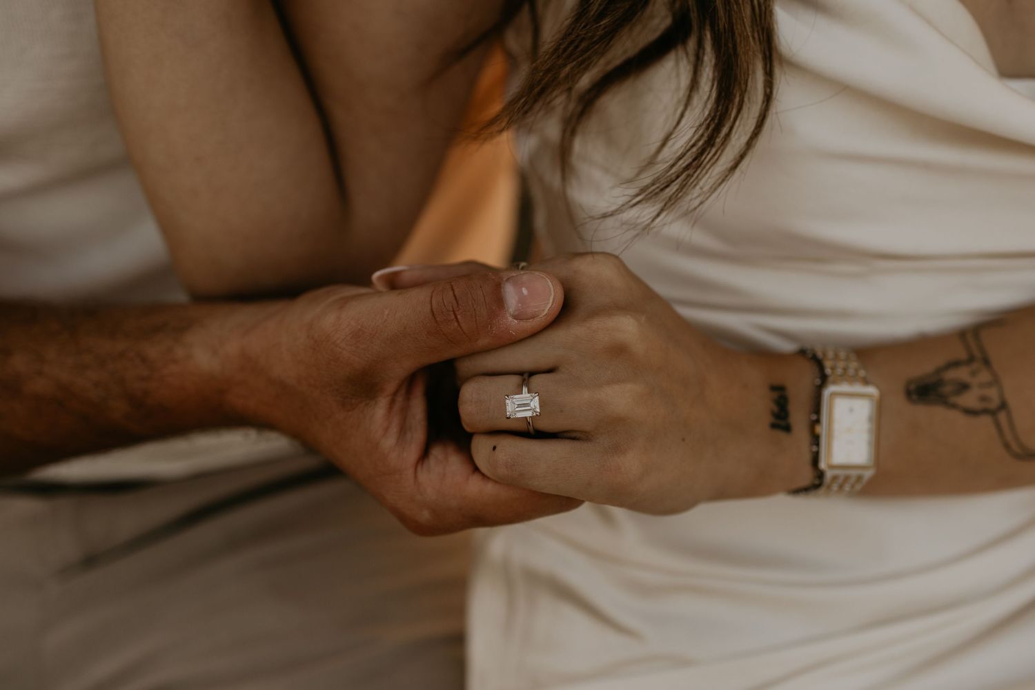 Close-up of hands showing engagement ring and tattoos against white fabric.