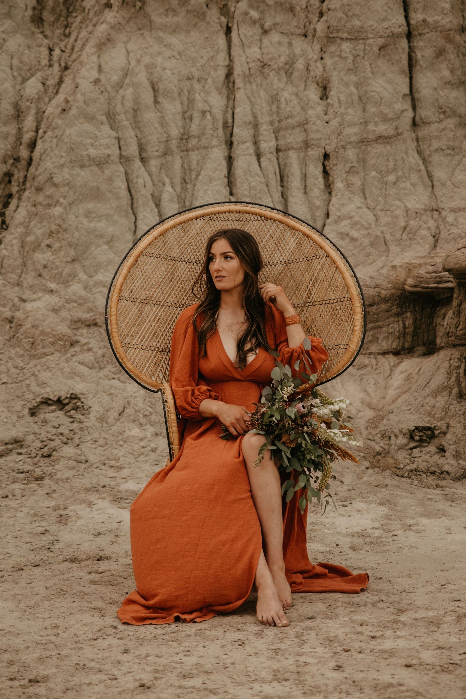 Person in orange dress sits in peacock chair holding flowers against clay cliff backdrop.