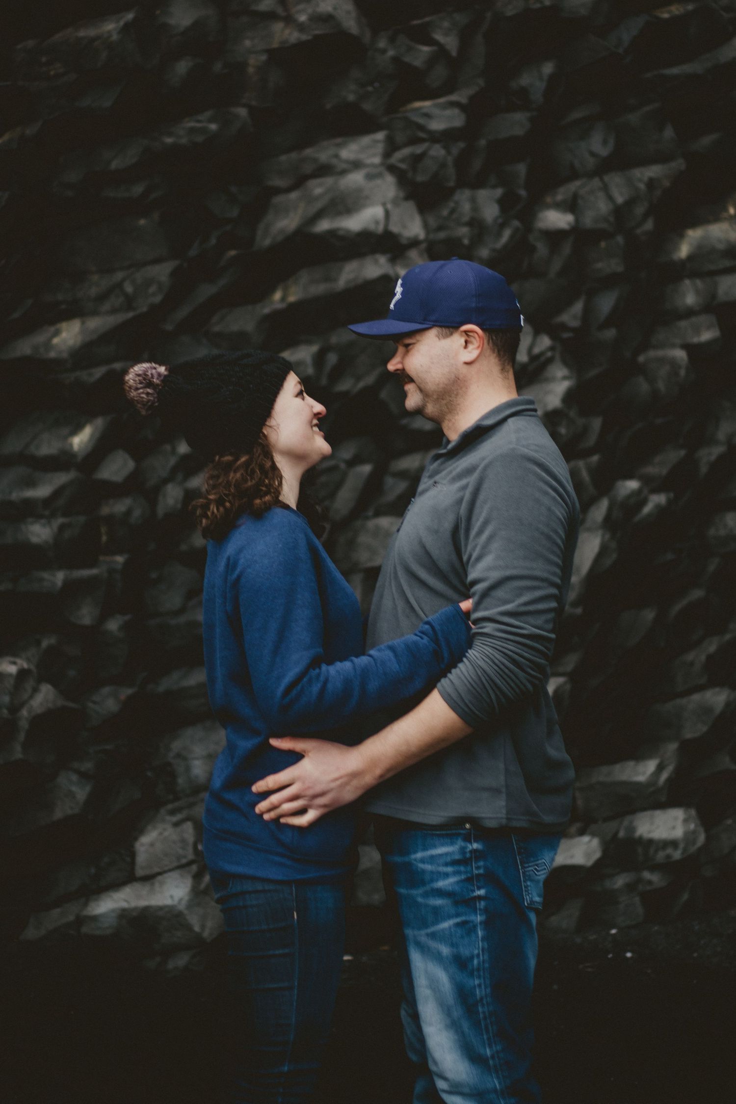 A romantic couple embrace against a dark stone wall outdoors, wearing coordinating blue and grey casual attire.