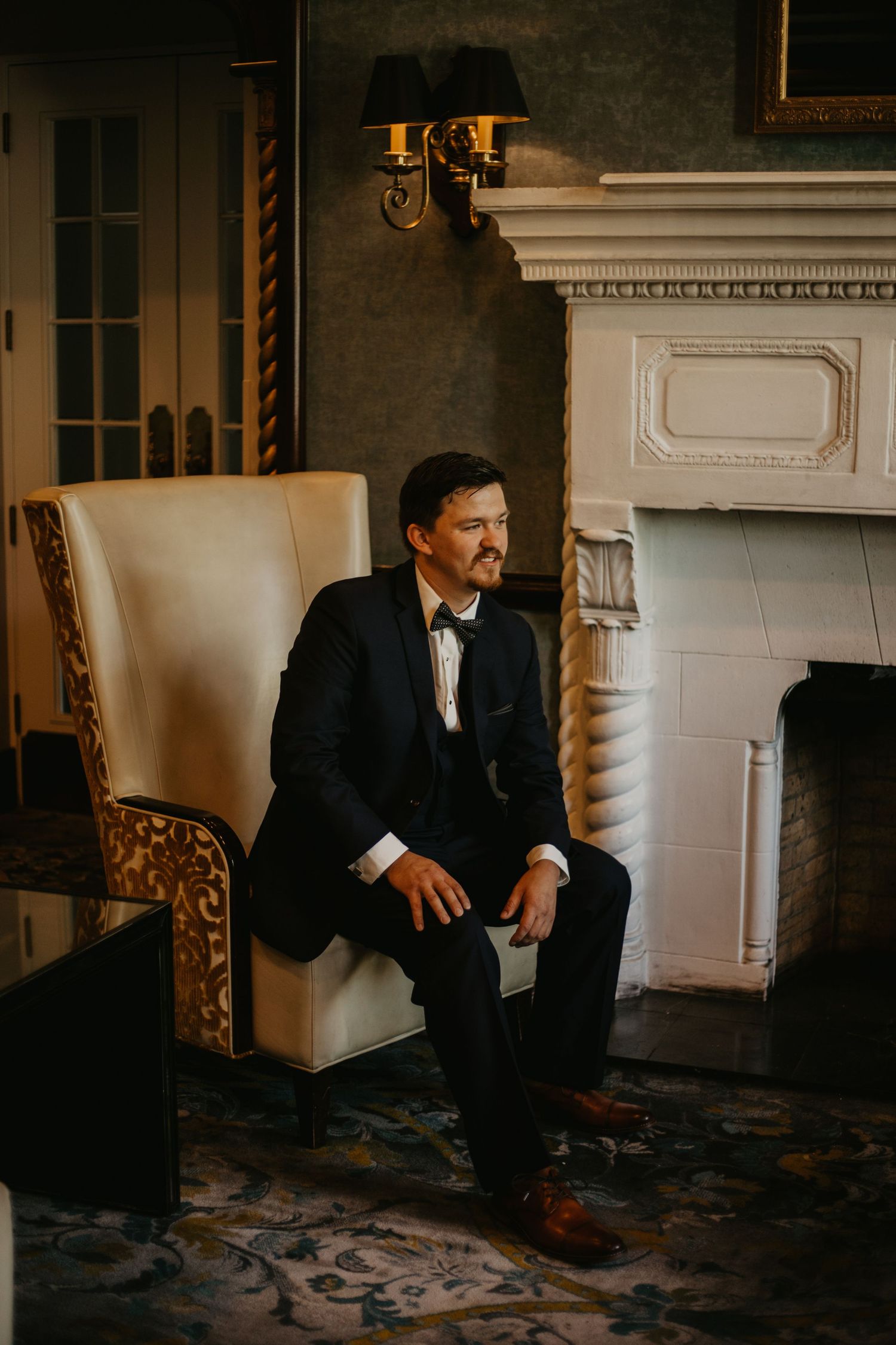 A contemplative moment in a formal suit beside an elegant white fireplace and upholstered chair.