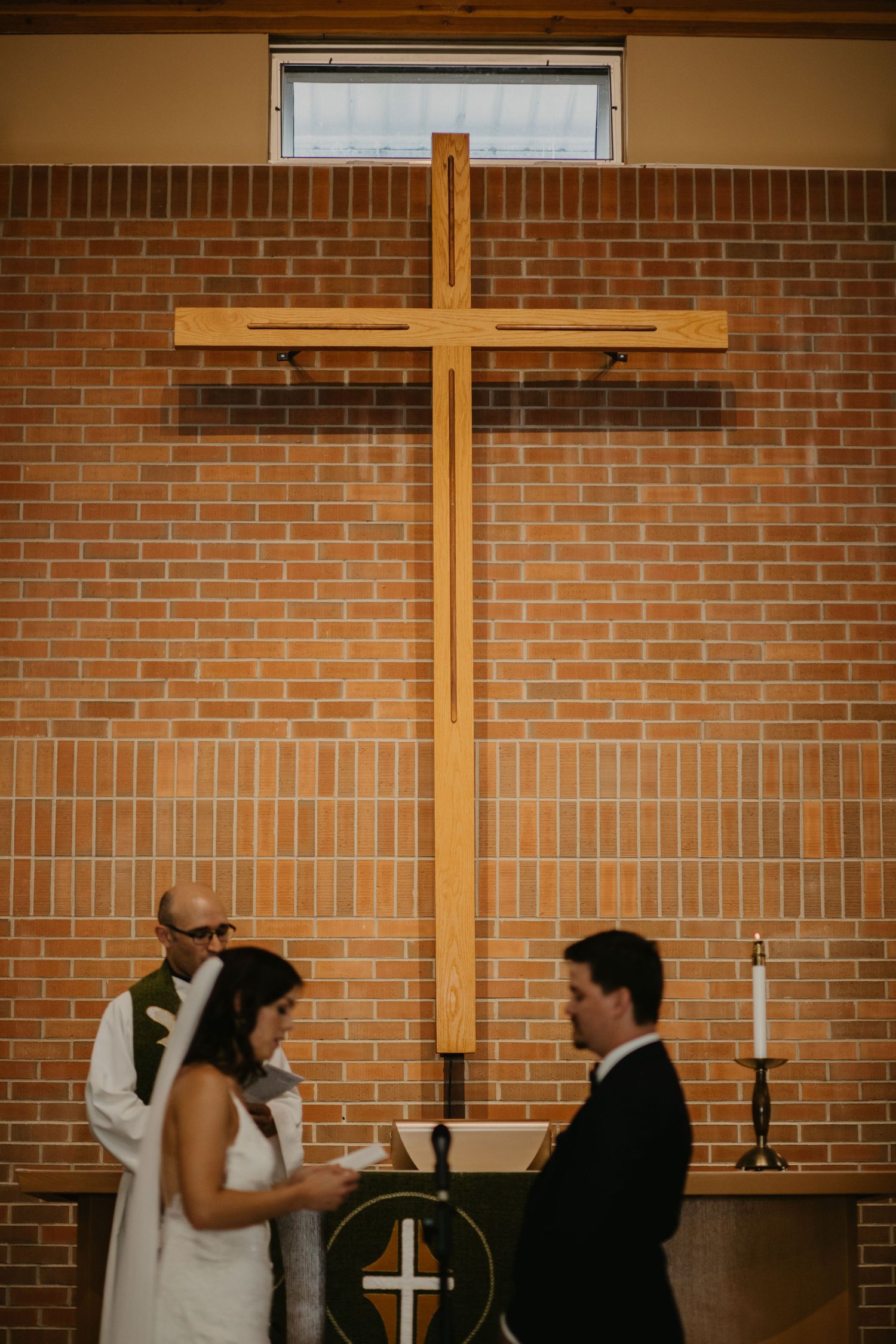A wedding ceremony takes place beneath a large wooden cross mounted on a brick wall.