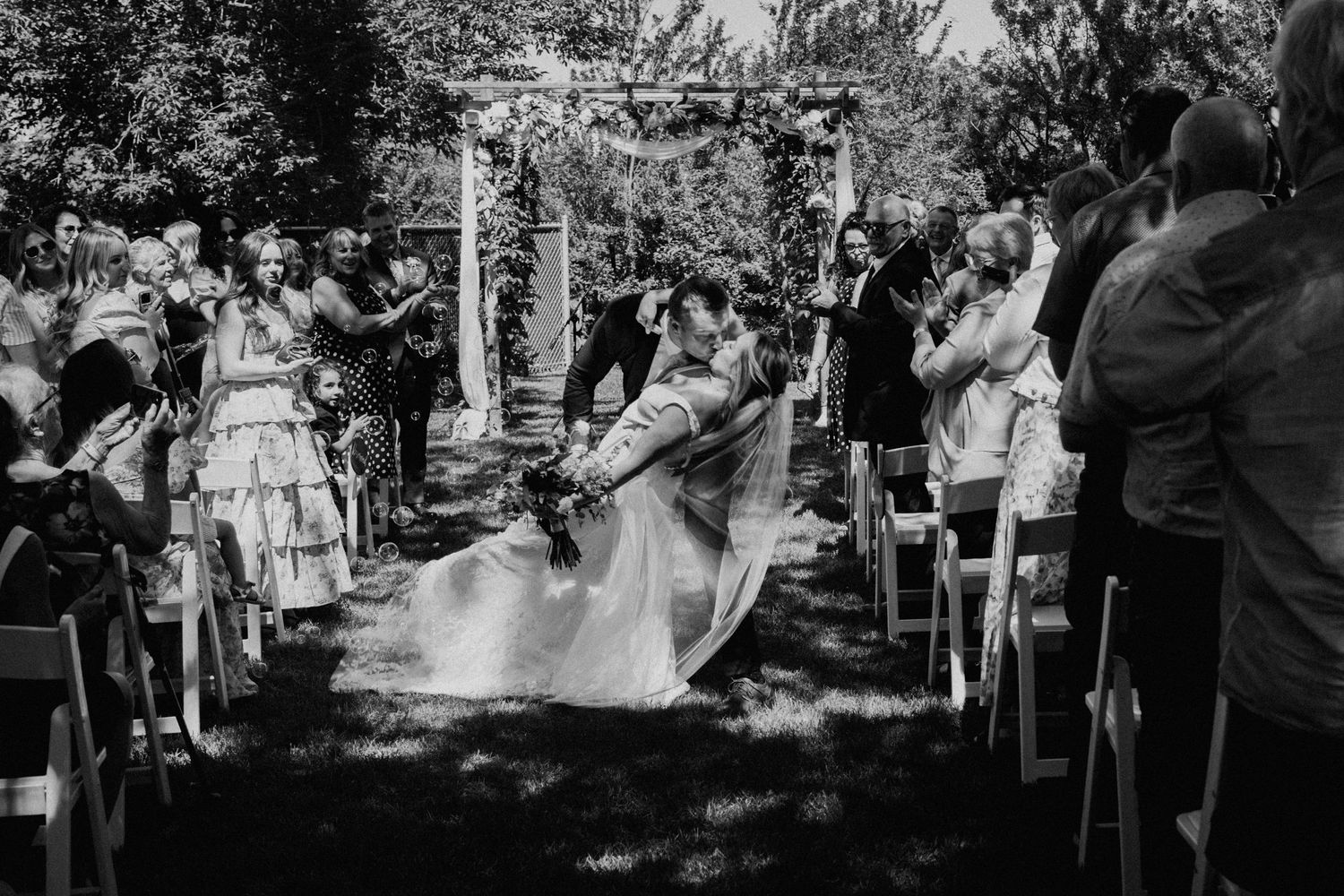 A black and white photo of an outdoor wedding ceremony with guests seated in rows of white chairs.