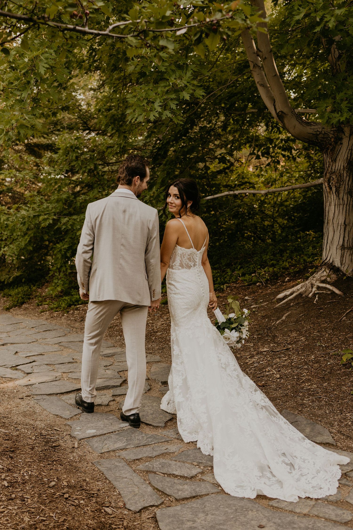A couple in wedding attire walking along a stone path surrounded by lush garden foliage.