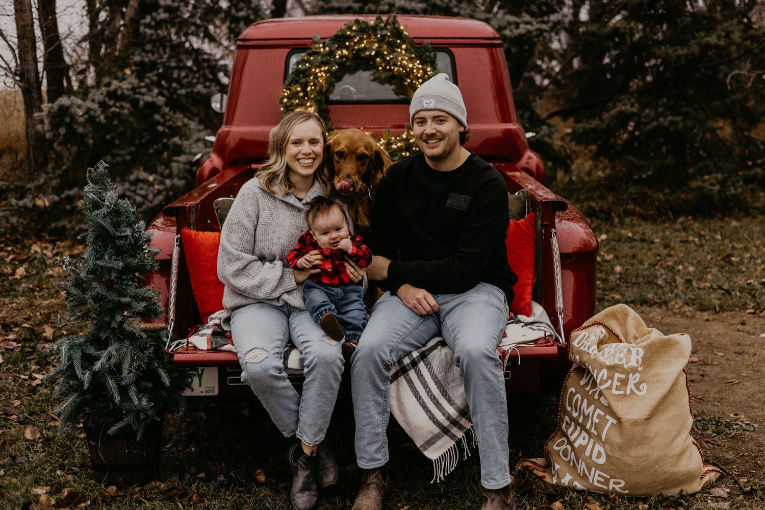 A family sits together in the bed of a vintage red pickup truck with blankets and bags.