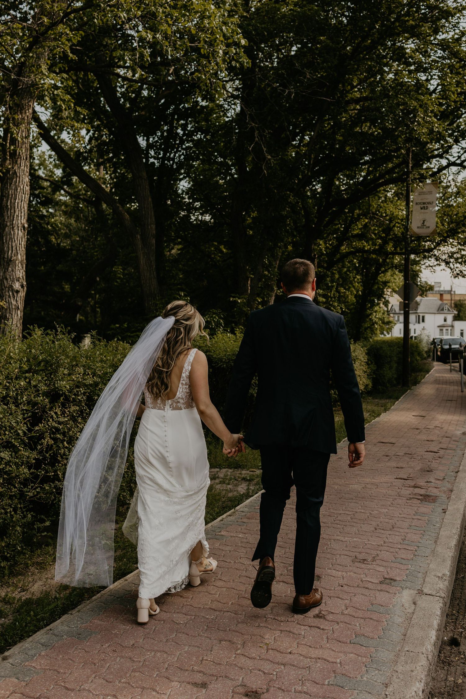 A bride and groom walk hand in hand down a tree-lined brick pathway on their wedding day.