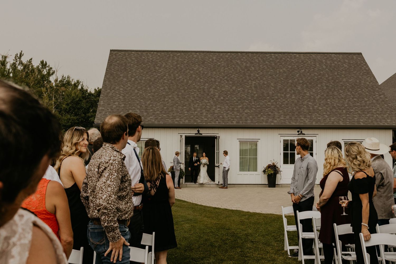 Guests gather outside a white farmhouse building for an outdoor wedding ceremony.