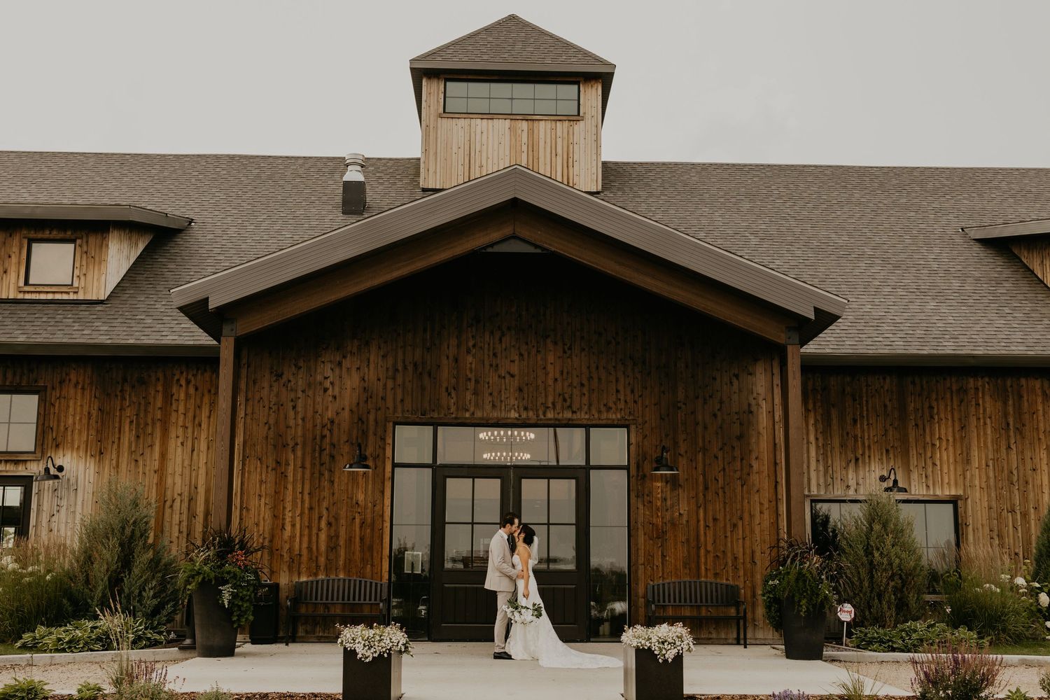 A rustic wooden barn wedding venue with a classic symmetrical entrance and potted plants along the walkway.