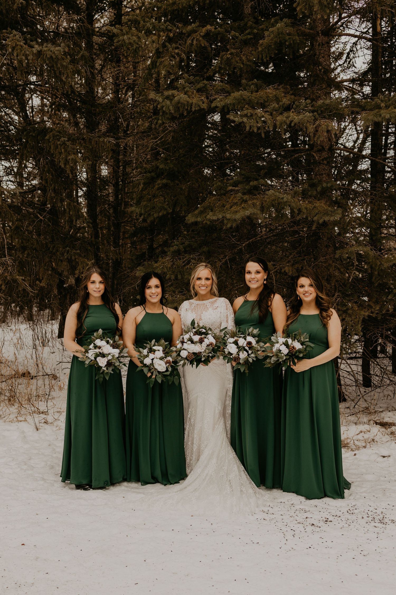 Bridal party in emerald green dresses and white gowns pose together against snowy forest backdrop.