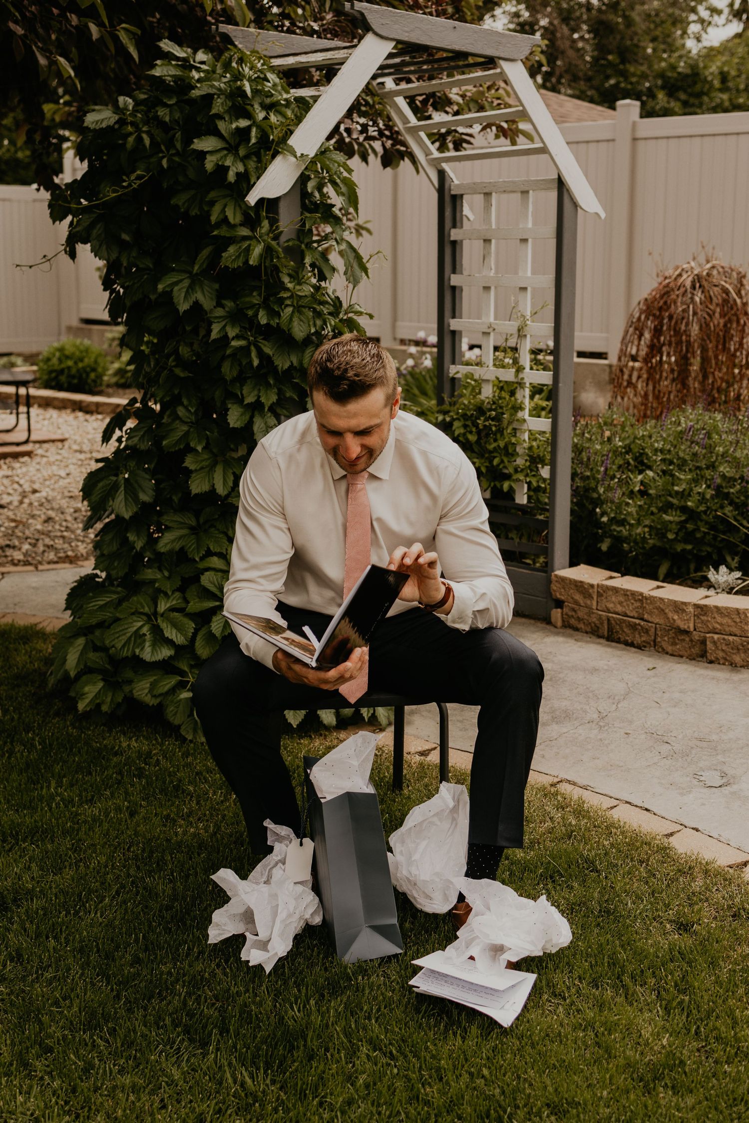 Groom reading notes while sitting in garden under white arbor surrounded by scattered papers.