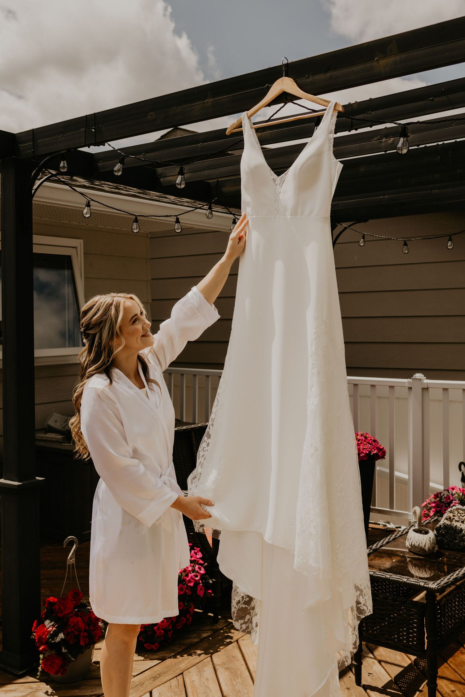 An elegant white wedding dress hanging on display outdoors under a wooden pergola.