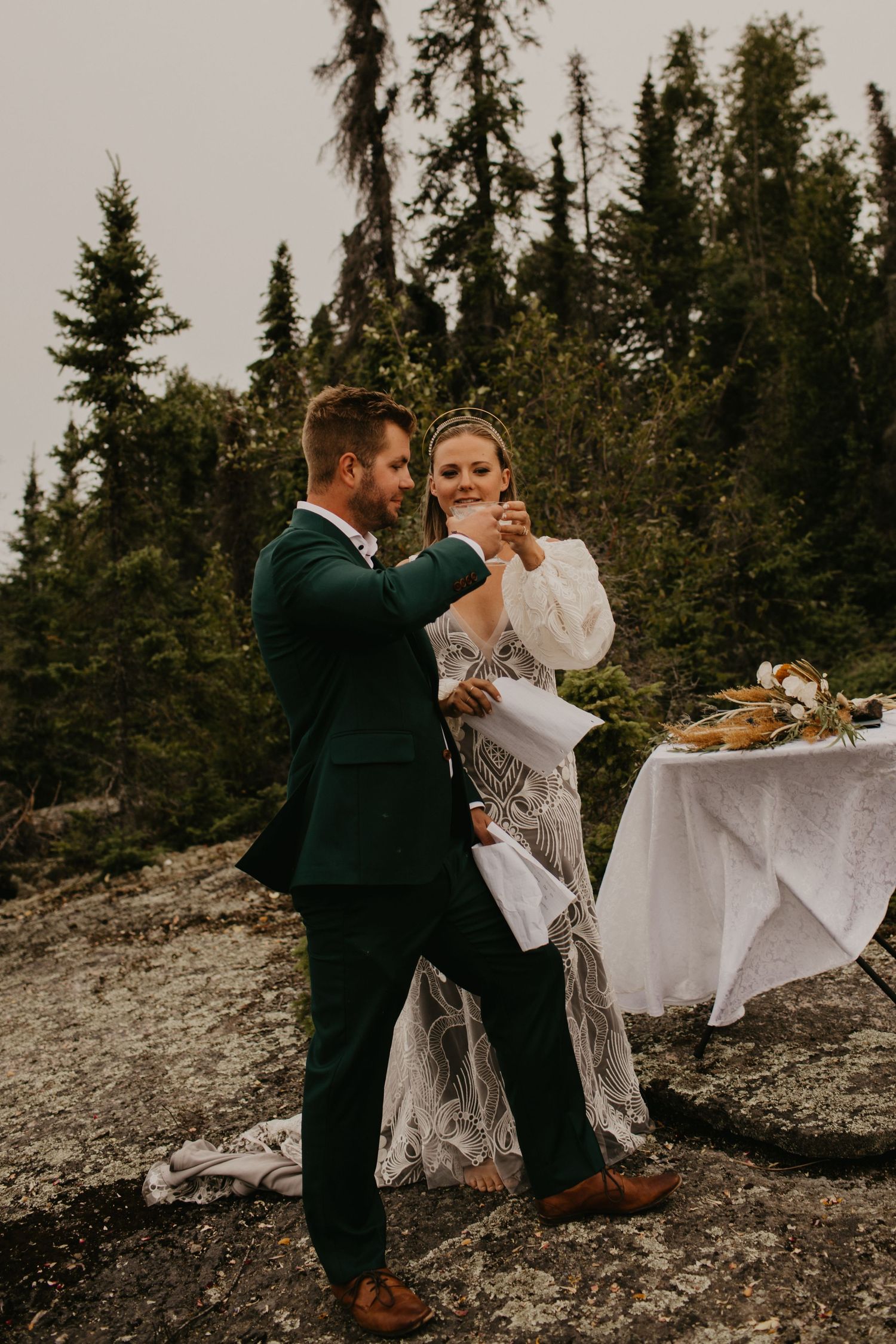 A couple shares a romantic dance moment by a small table on a rocky cliff surrounded by evergreen trees.