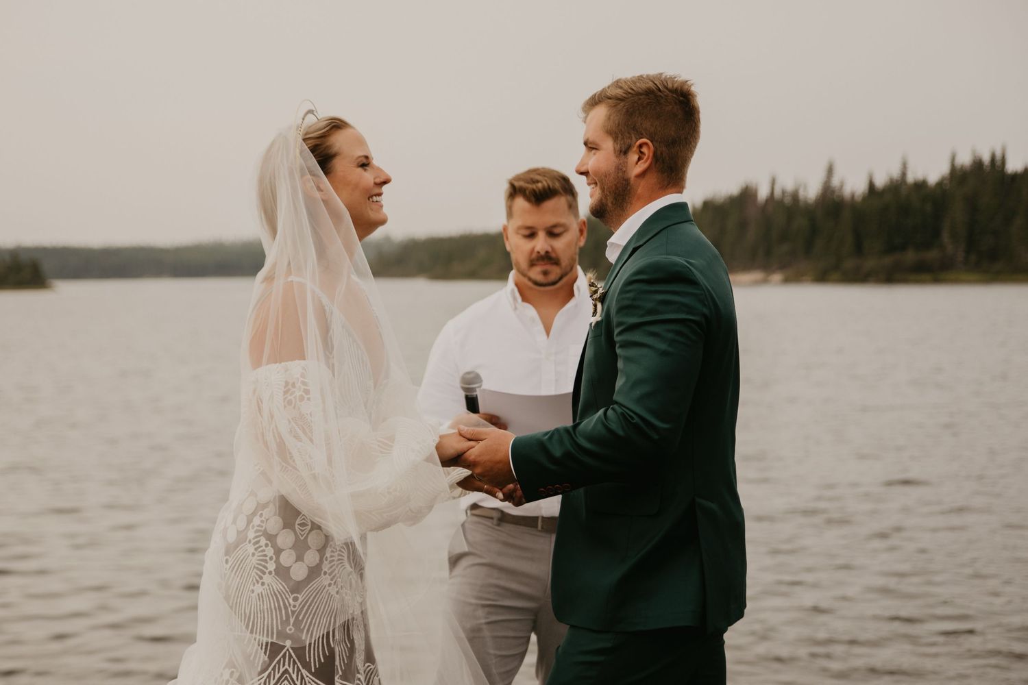 Wedding ceremony in progress on shoreline with forest and lake backdrop.