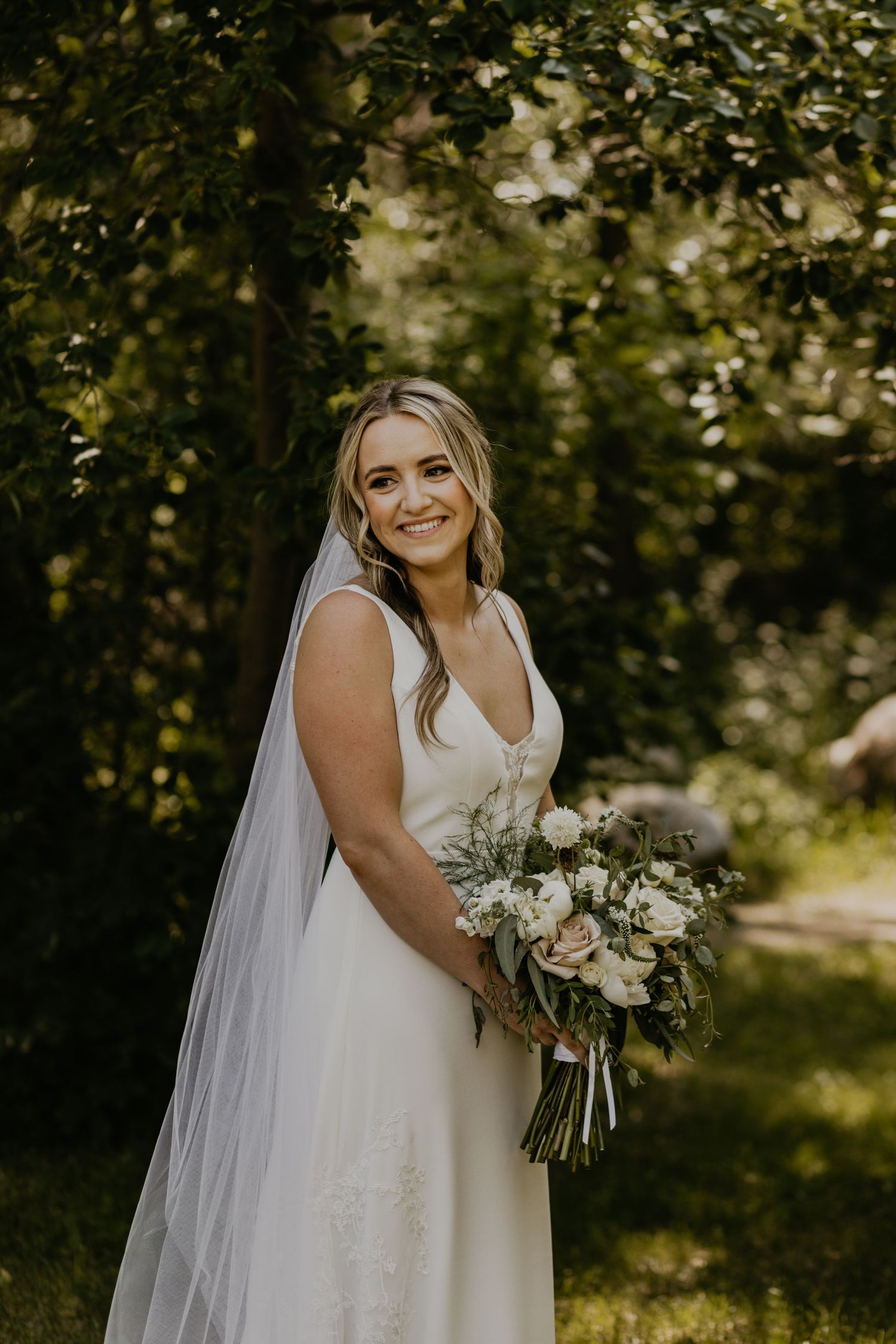 Bride in elegant white dress holds bouquet while posing in sunlit garden.