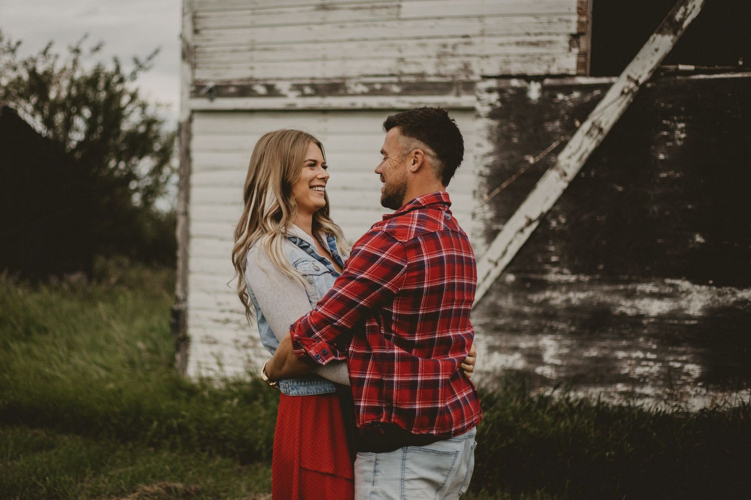 A couple embraces in front of a weathered white barn in vintage style.