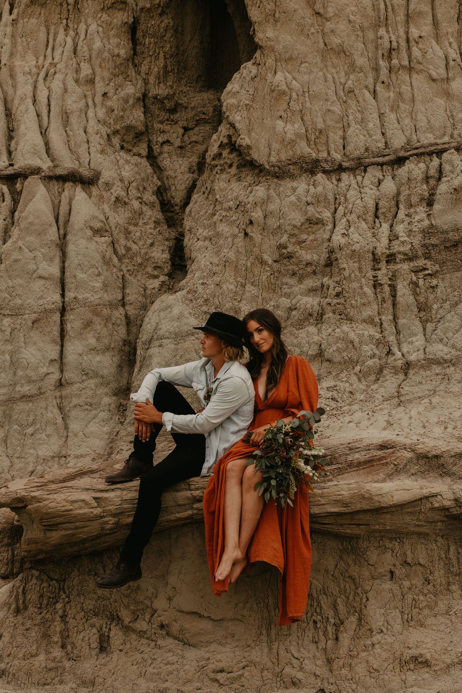 A stylish couple sits on a ledge against towering clay cliffs in desert terrain.