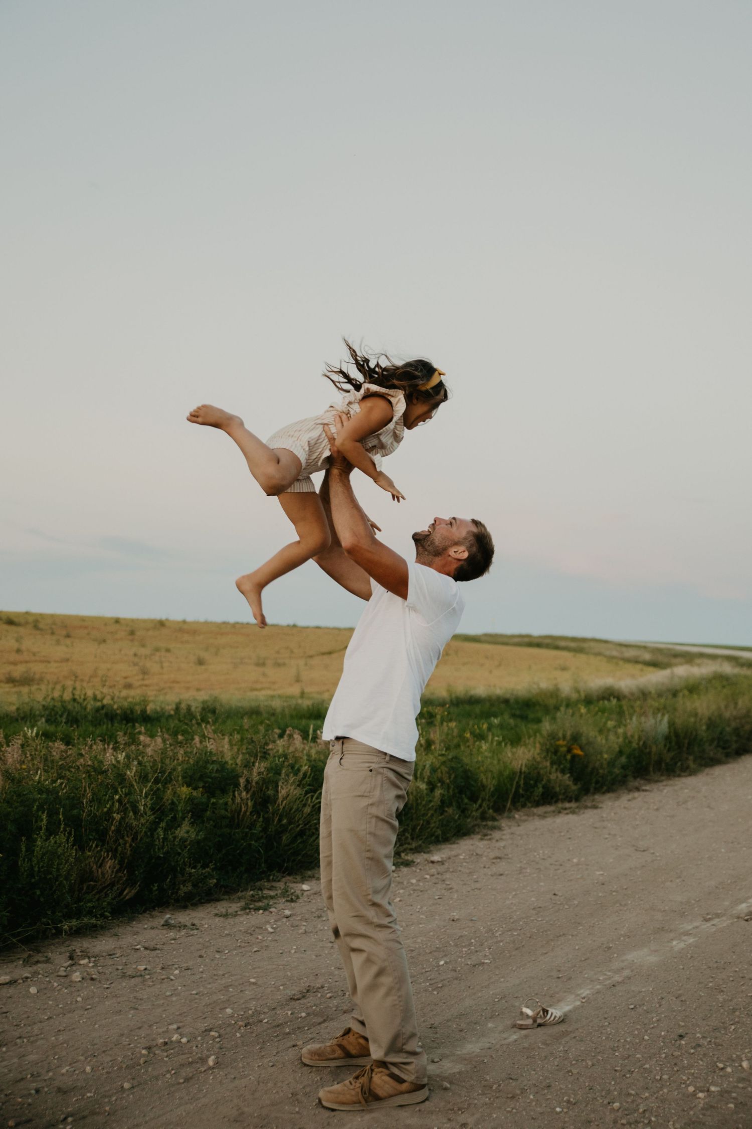 Playful moment captured on dirt path as figure lifts child high in countryside setting at dusk.