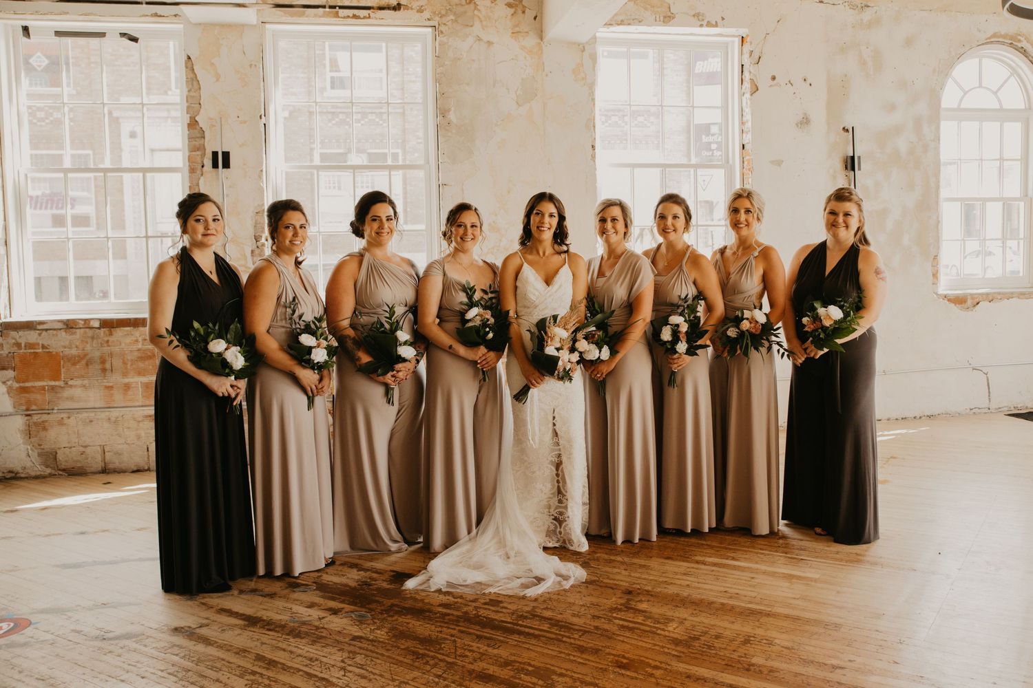Wedding party in coordinated neutral gowns stands in line against white brick walls.