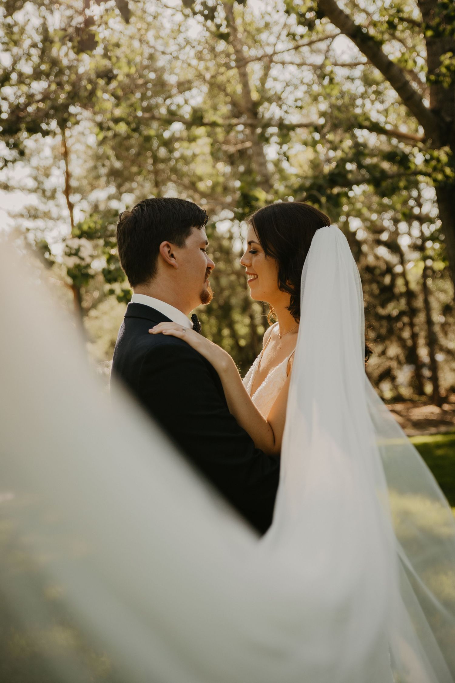 Intimate wedding portrait through sheer veil in sunlit wooded setting.