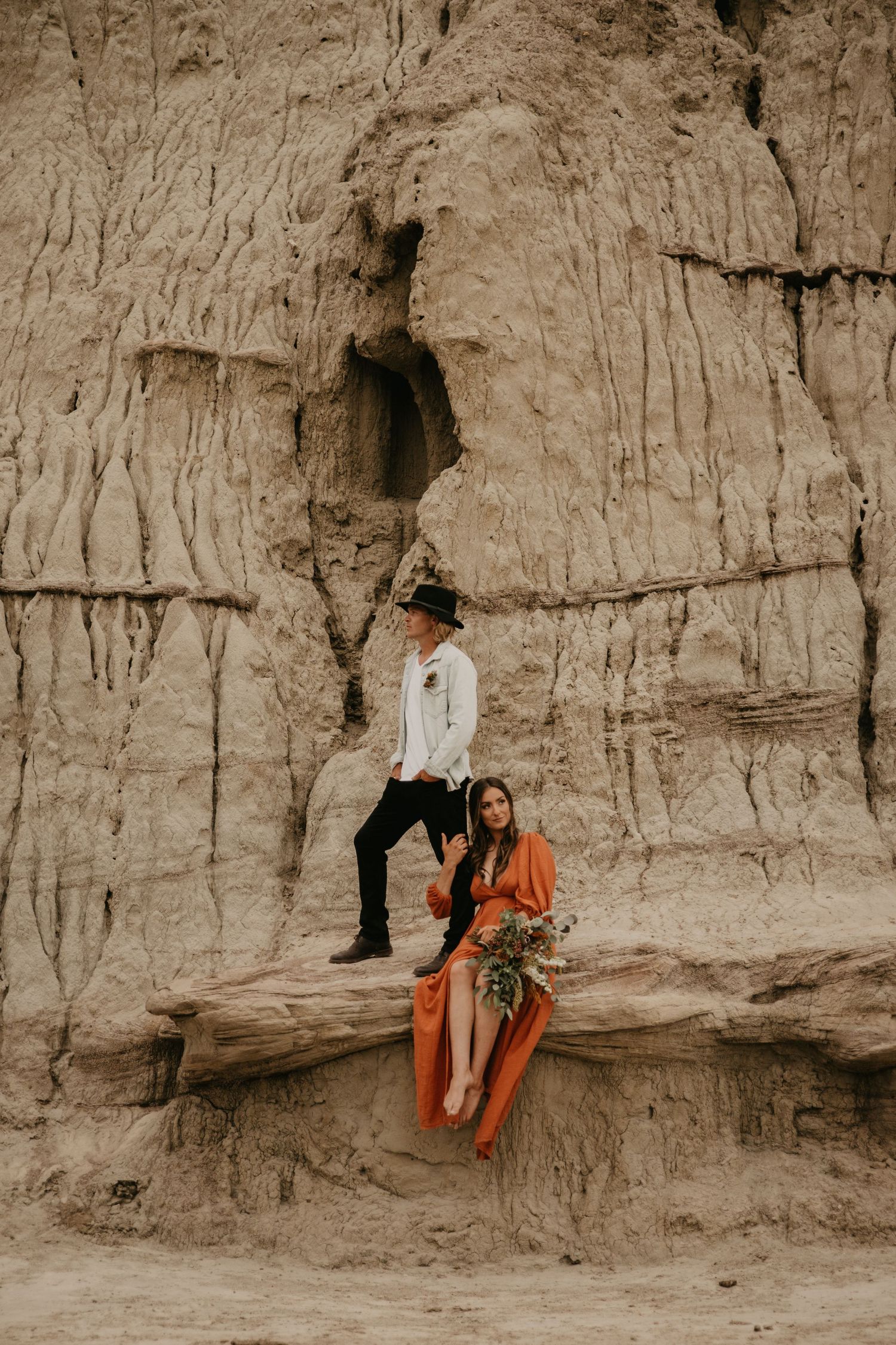 Two people pose dramatically against rugged clay cliffs in contrasting white and orange outfits.