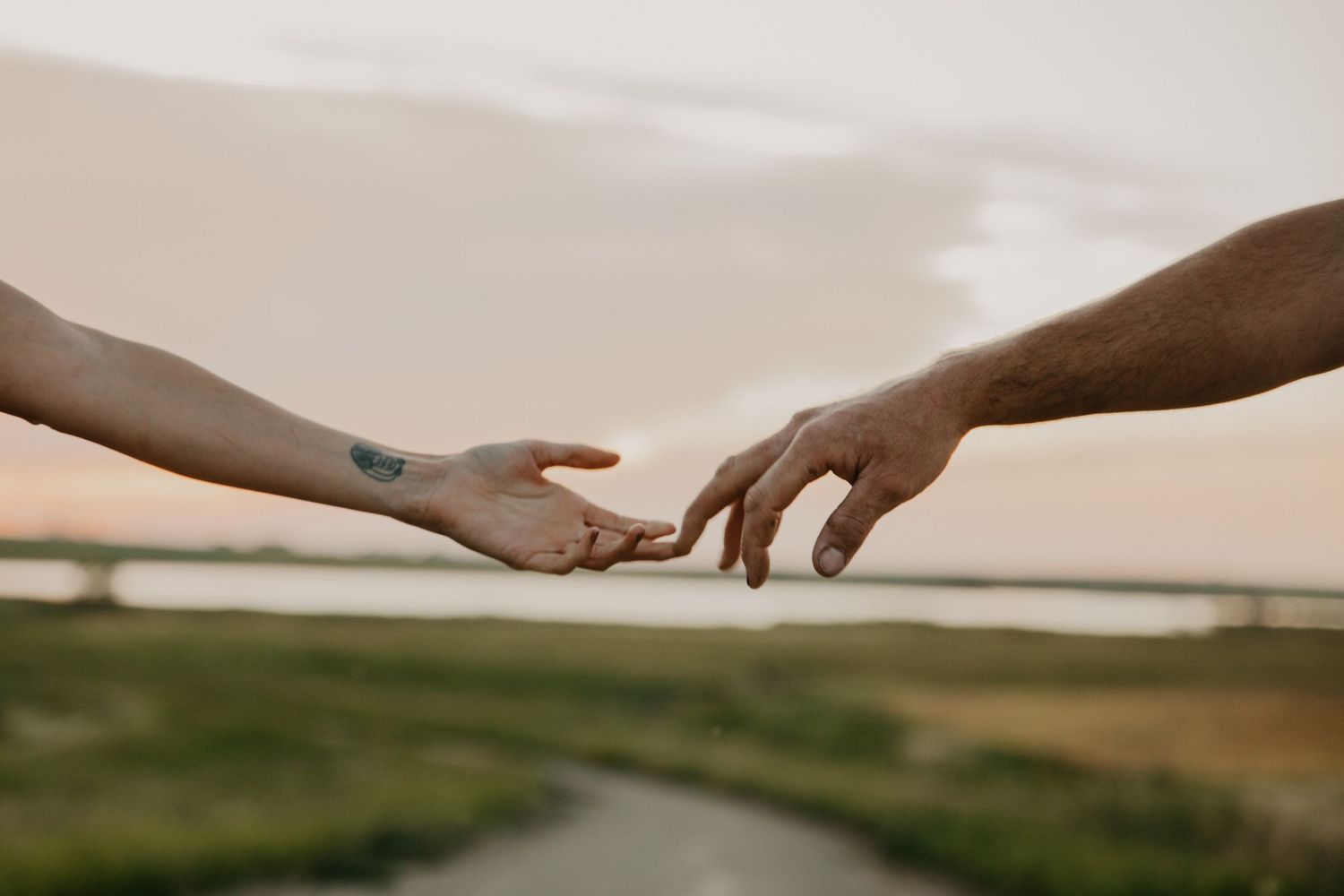 Two hands reaching toward each other against sunset sky over rural landscape.