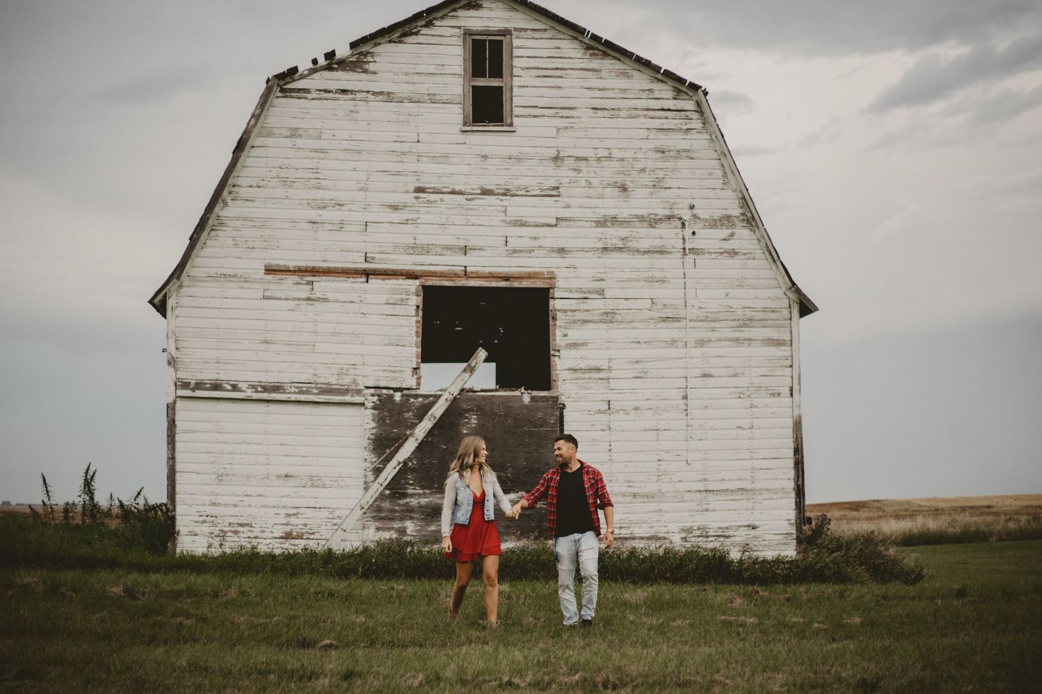 A couple stands holding hands in front of an old white barn structure.