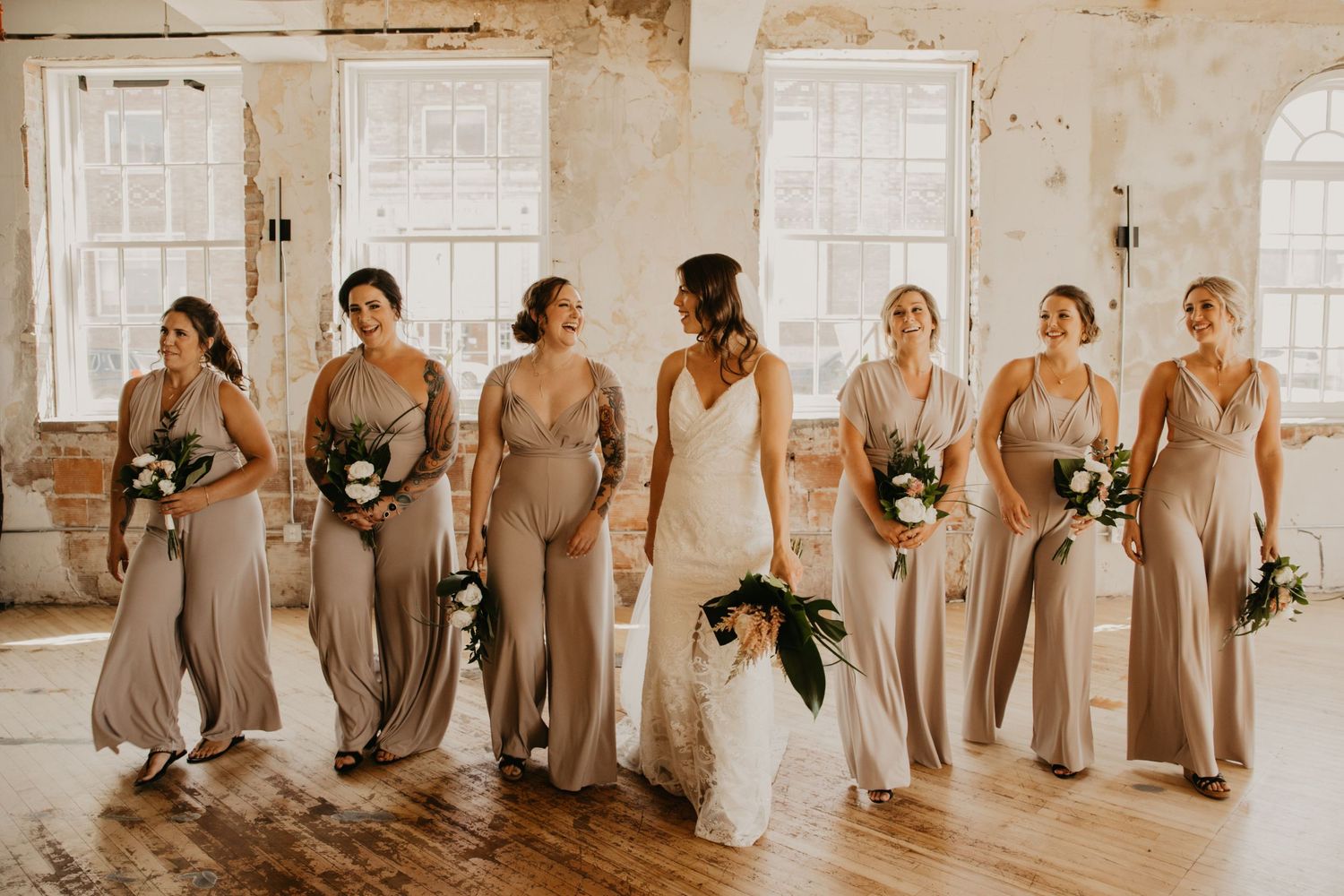 Bridesmaids in champagne colored dresses pose with bride in white gown in rustic venue.
