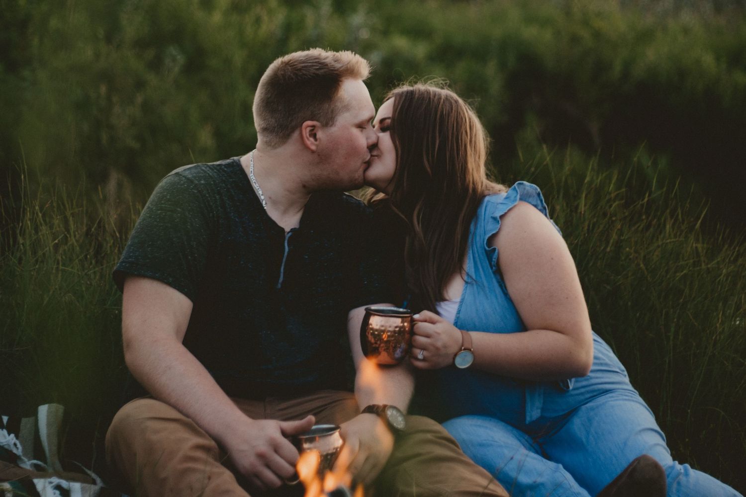 Couple sharing drinks and kisses by a cozy campfire at twilight.