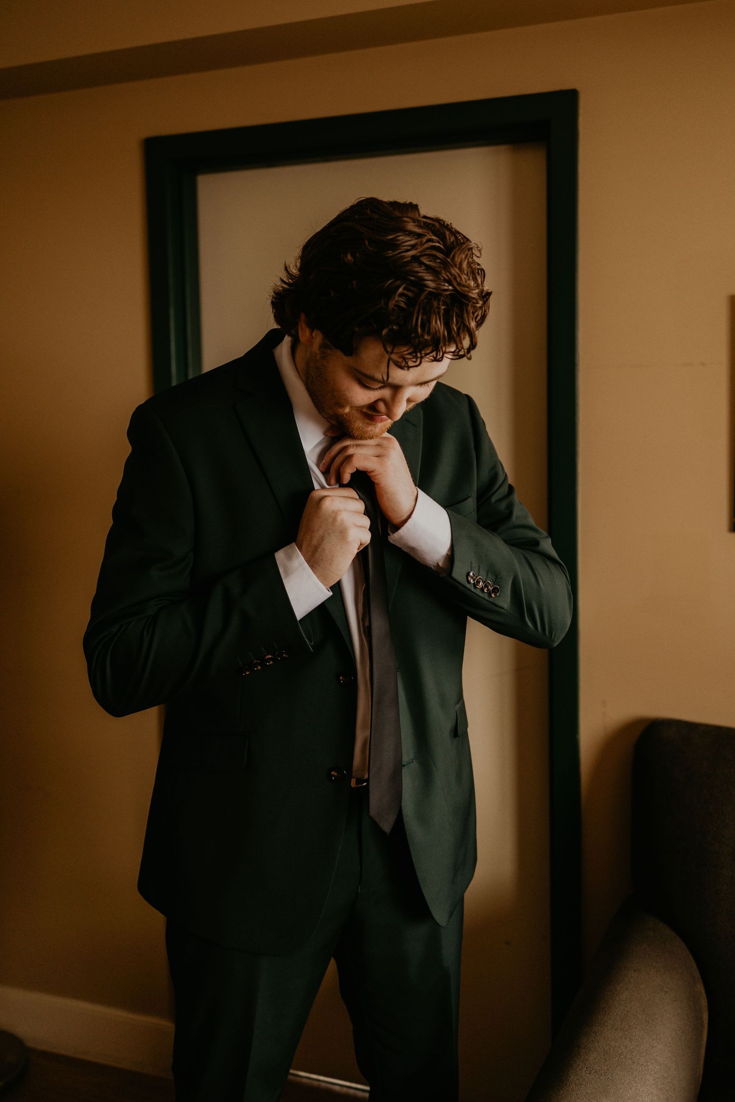 Person in dark suit adjusting tie while getting ready in warm-toned indoor setting.