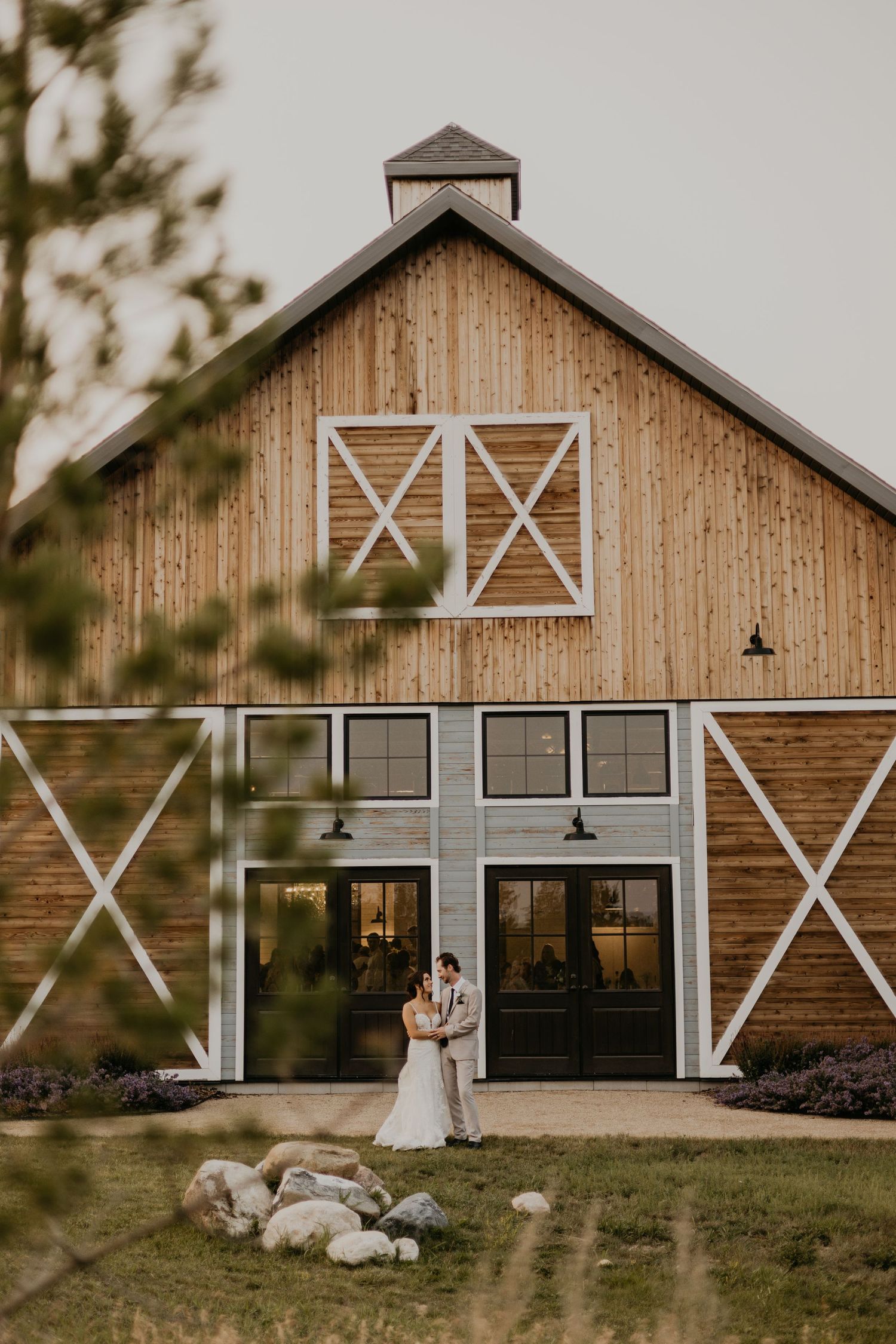 Modern barn wedding venue with couple standing in front of large wooden doors at sunset.