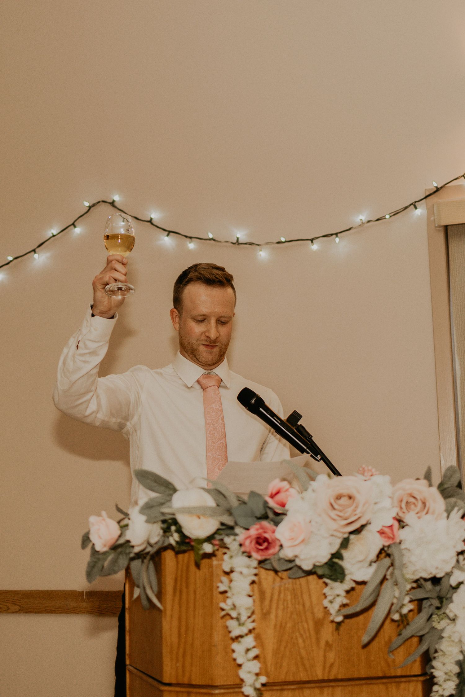 A wedding toast speech being given at a reception decorated with string lights and pink roses.