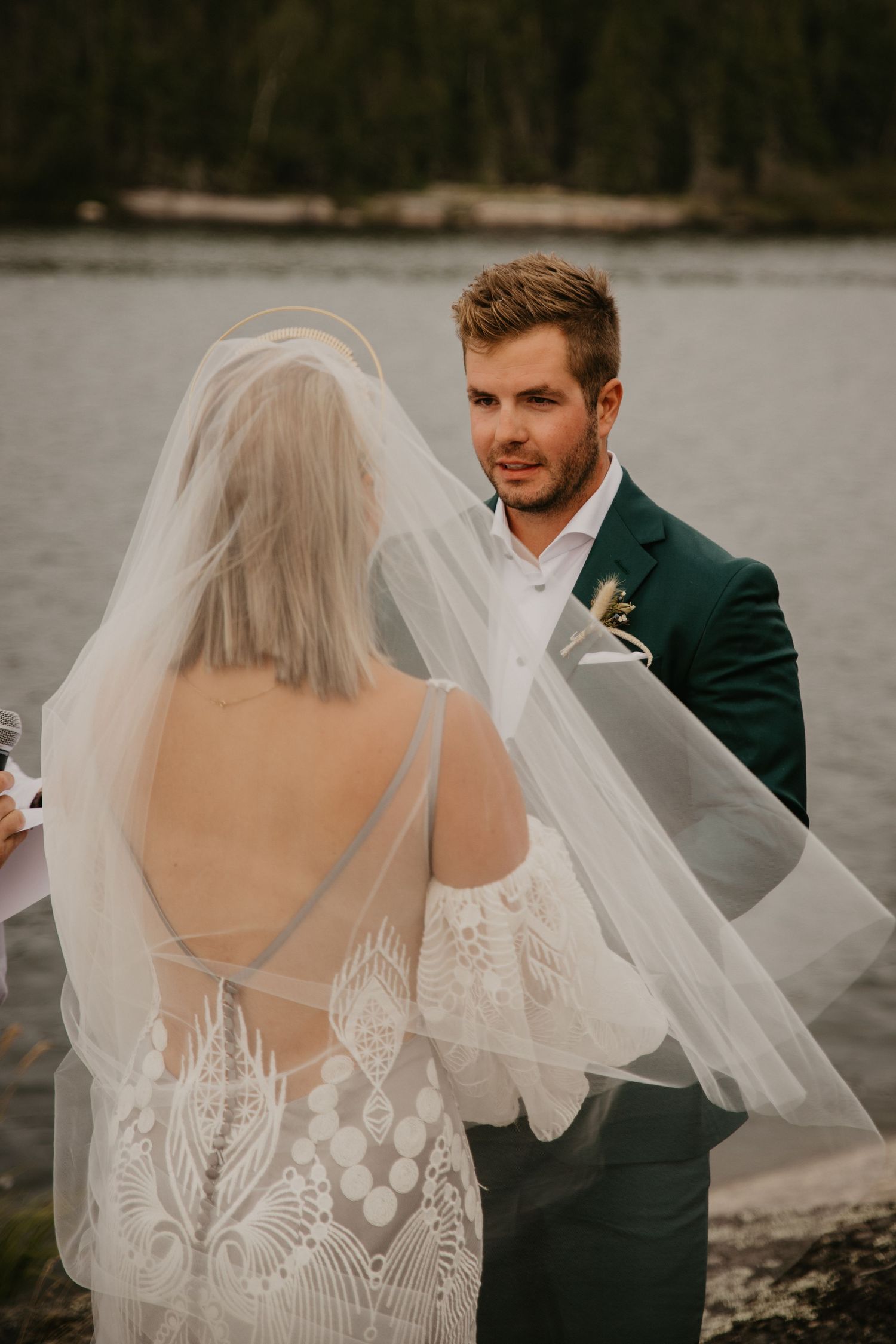 A couple shares an intimate wedding moment by a lake with the bride wearing a detailed lace dress and flowing veil.