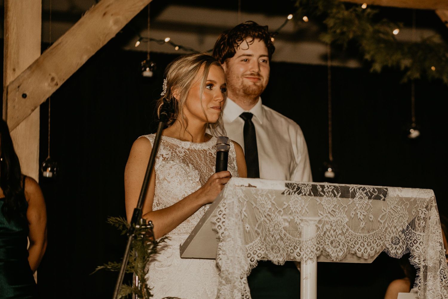 Wedding couple delivers speeches at a rustic reception venue with lace-covered podium.
