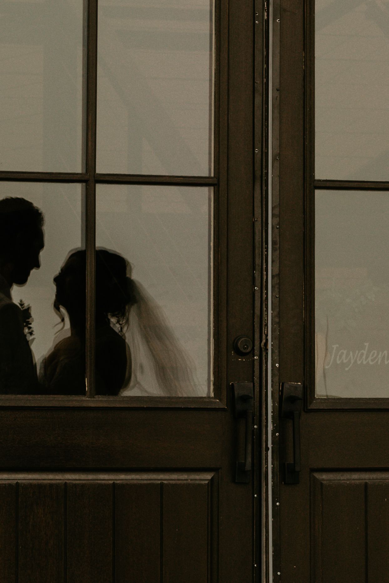 Wedding party in beige suits and black dresses poses formally outside a rustic wooden venue.