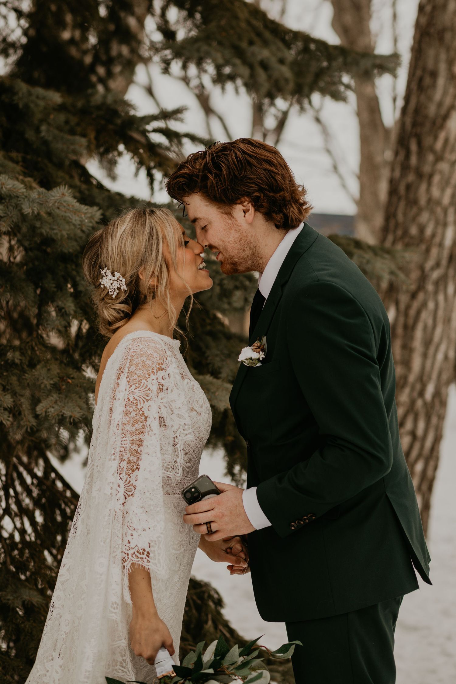 An intimate wedding moment captured between a couple beneath pine trees in winter.